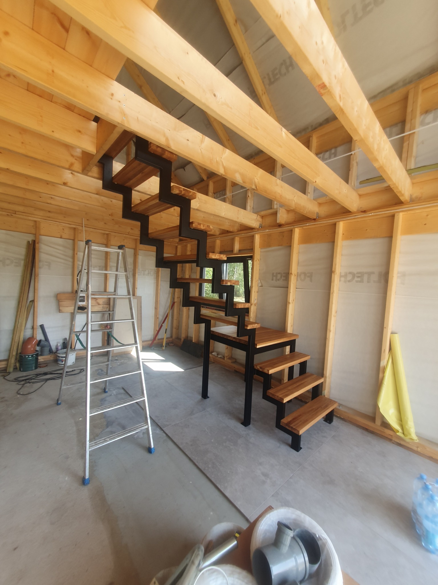 Schody o konstrukcji metalowej z drewnianymi stopniami, zamontowane w surowym wnętrzu z drewnianą konstrukcją dachu, widoczna drabina i materiały budowlane.