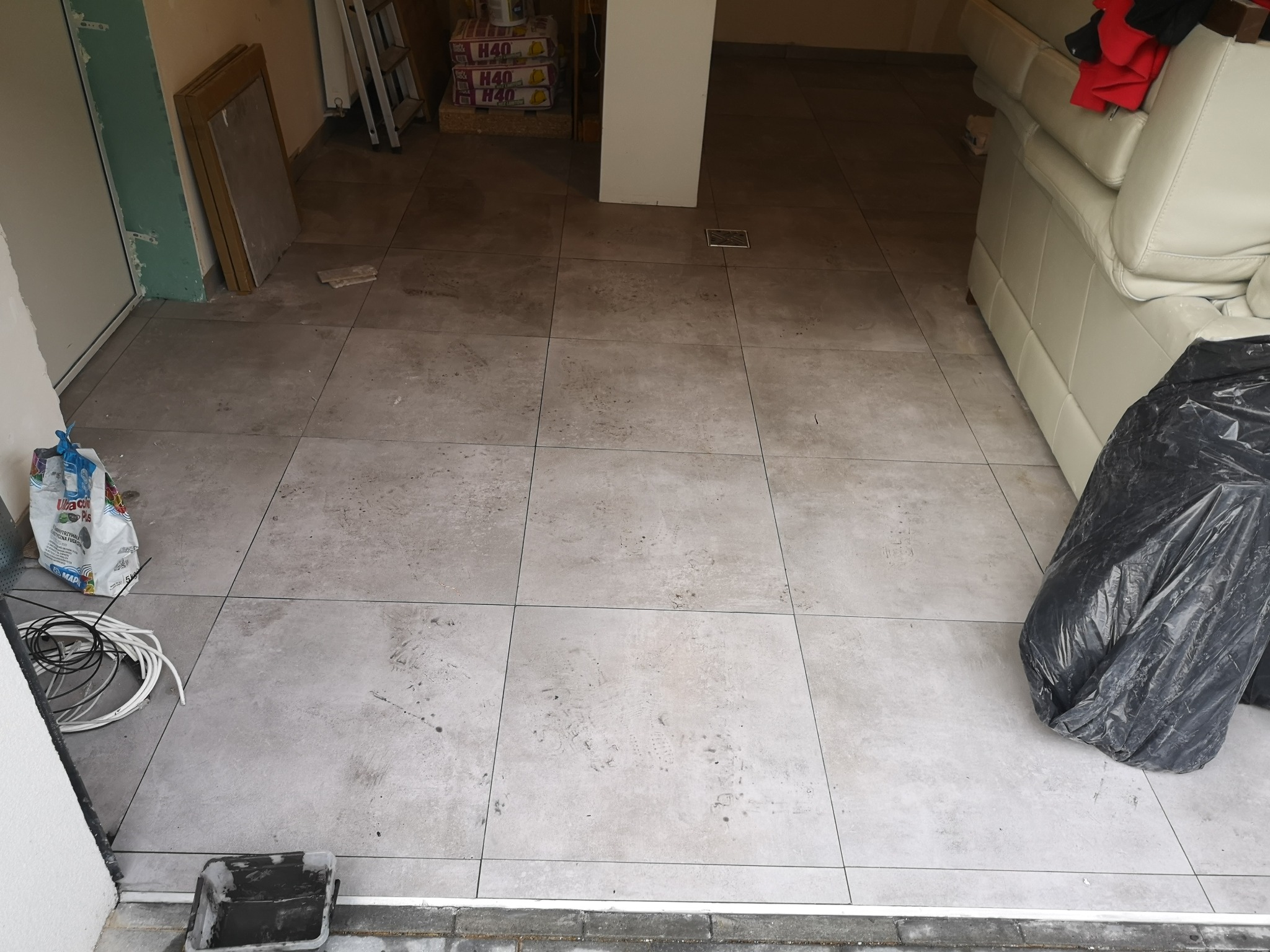 Ułożone szare płytki podłogowe w trakcie prac wykończeniowych w Warszawie, widoczny worek z odpadami budowlanymi, wiadro po zaprawie i zwój kabli.