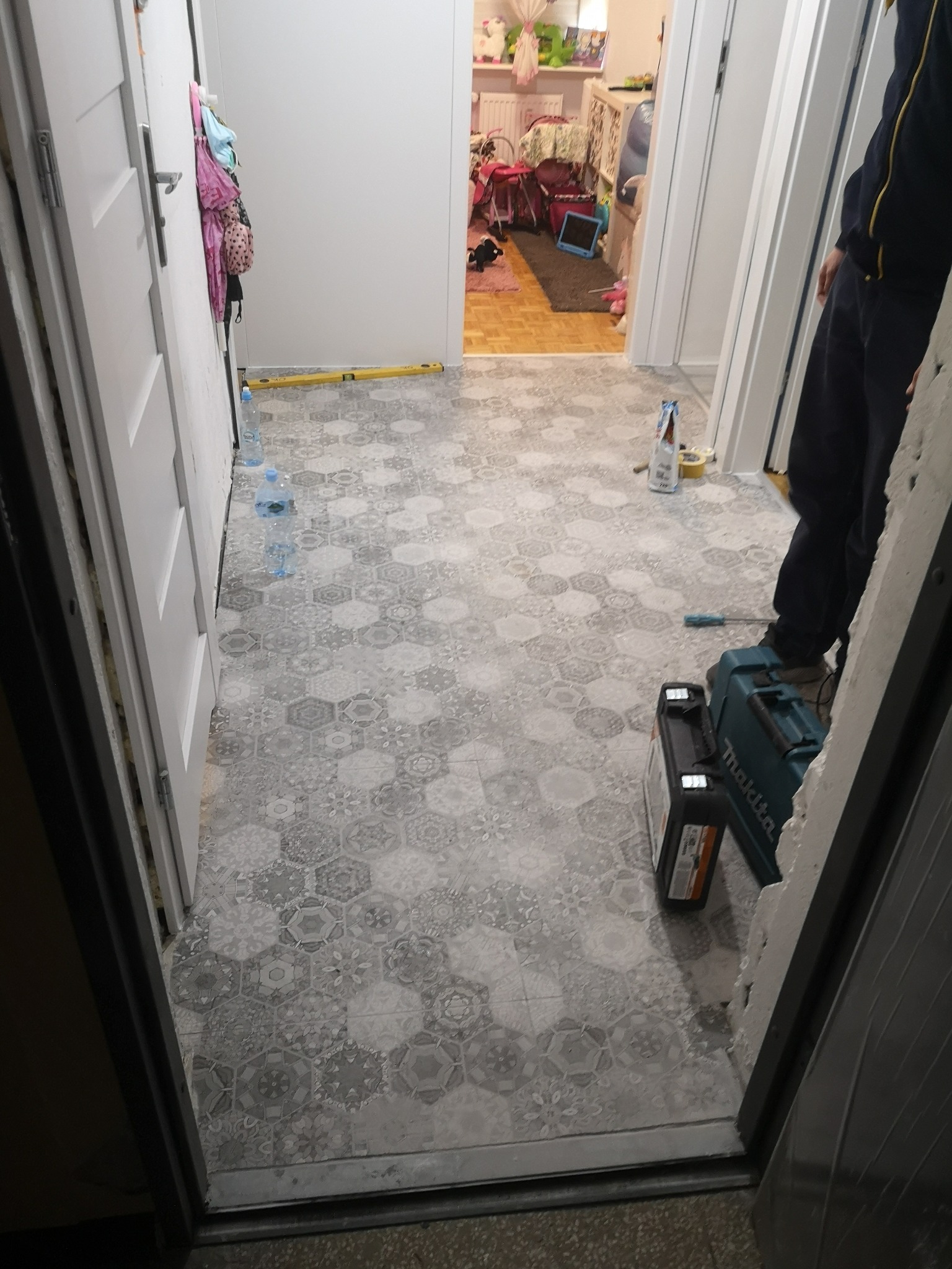 Układanie heksagonalnych płytek podłogowych w korytarzu, widoczna skrzynka narzędziowa Makita, poziomica, butelki z wodą i częściowo postać instalatora.
