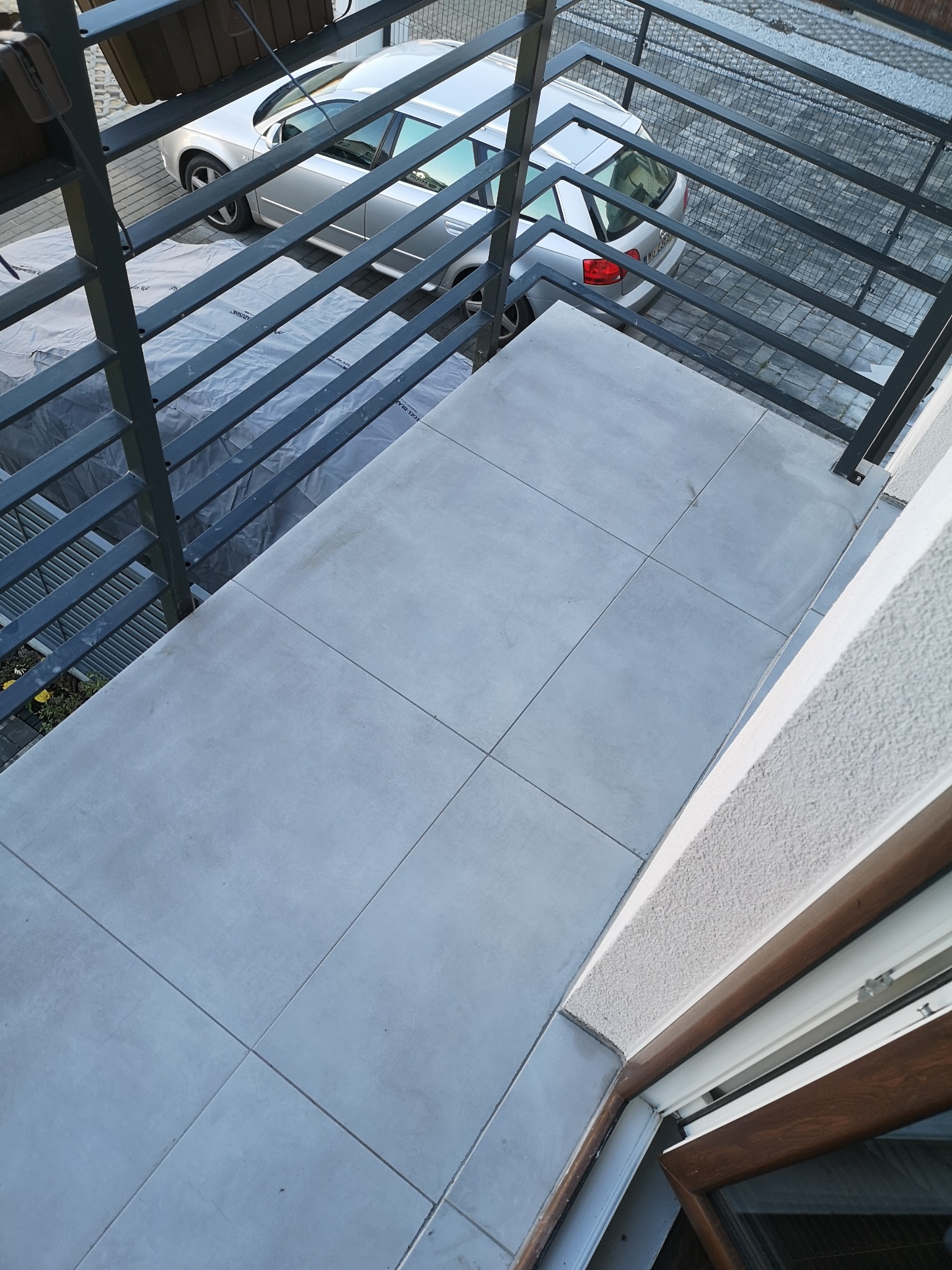 Jasnoszara, nowo położona podłoga z płytek na balkonie, widok z góry, z fragmentem szarej barierki i zaparkowanym srebrnym samochodem w tle.