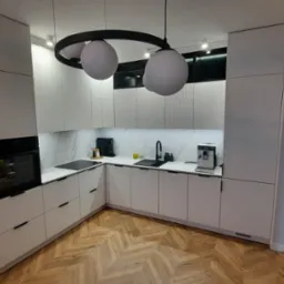 Nowoczesna kuchnia z białymi frontami, czarnymi akcentami i parkietem w jodełkę, oświetlona lampą z trzema kulami.