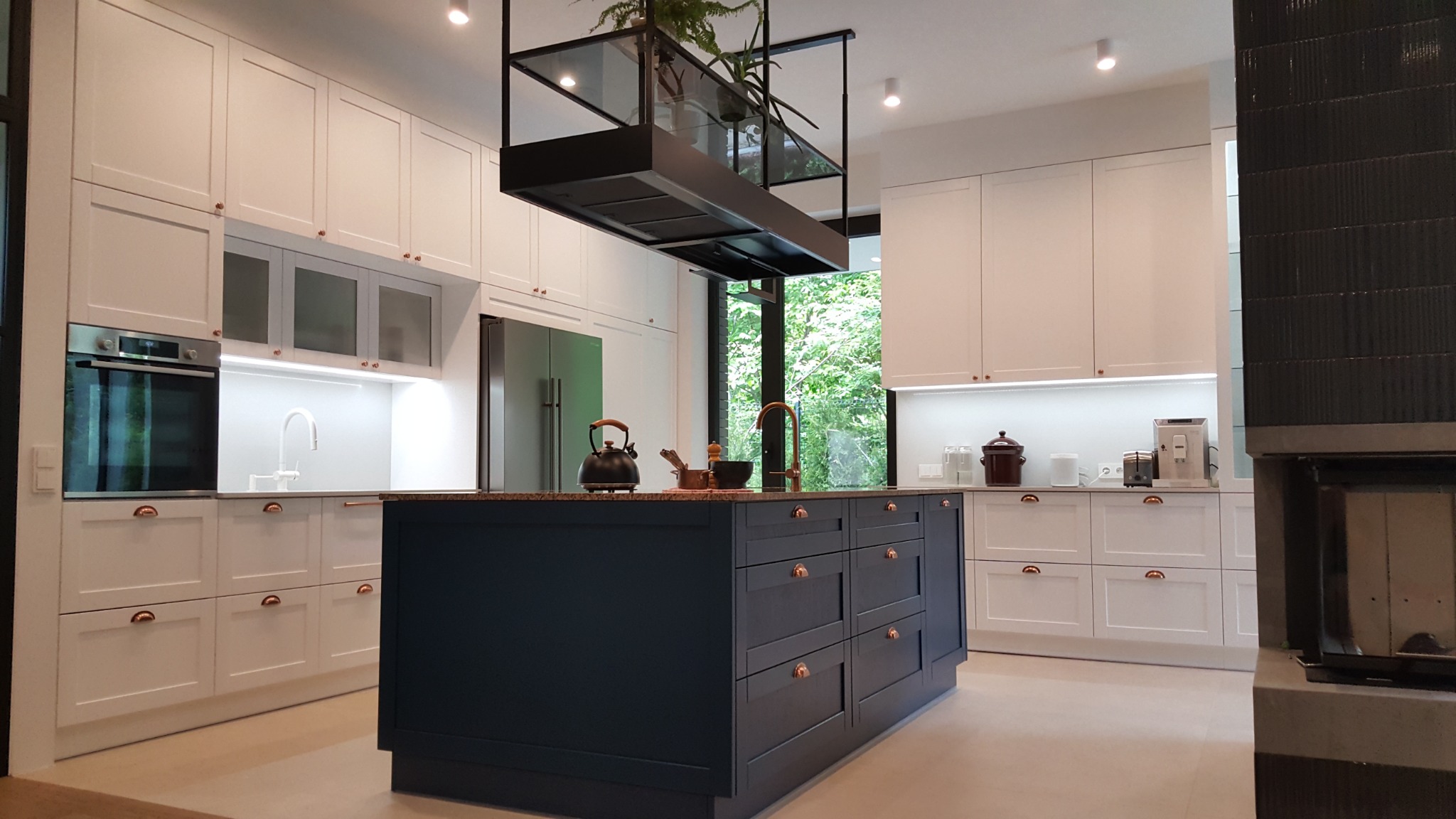 Nowoczesna kuchnia z białymi szafkami, wyspą w kolorze granatowym z miedzianymi uchwytami i wiszącym okapem z roślinami.