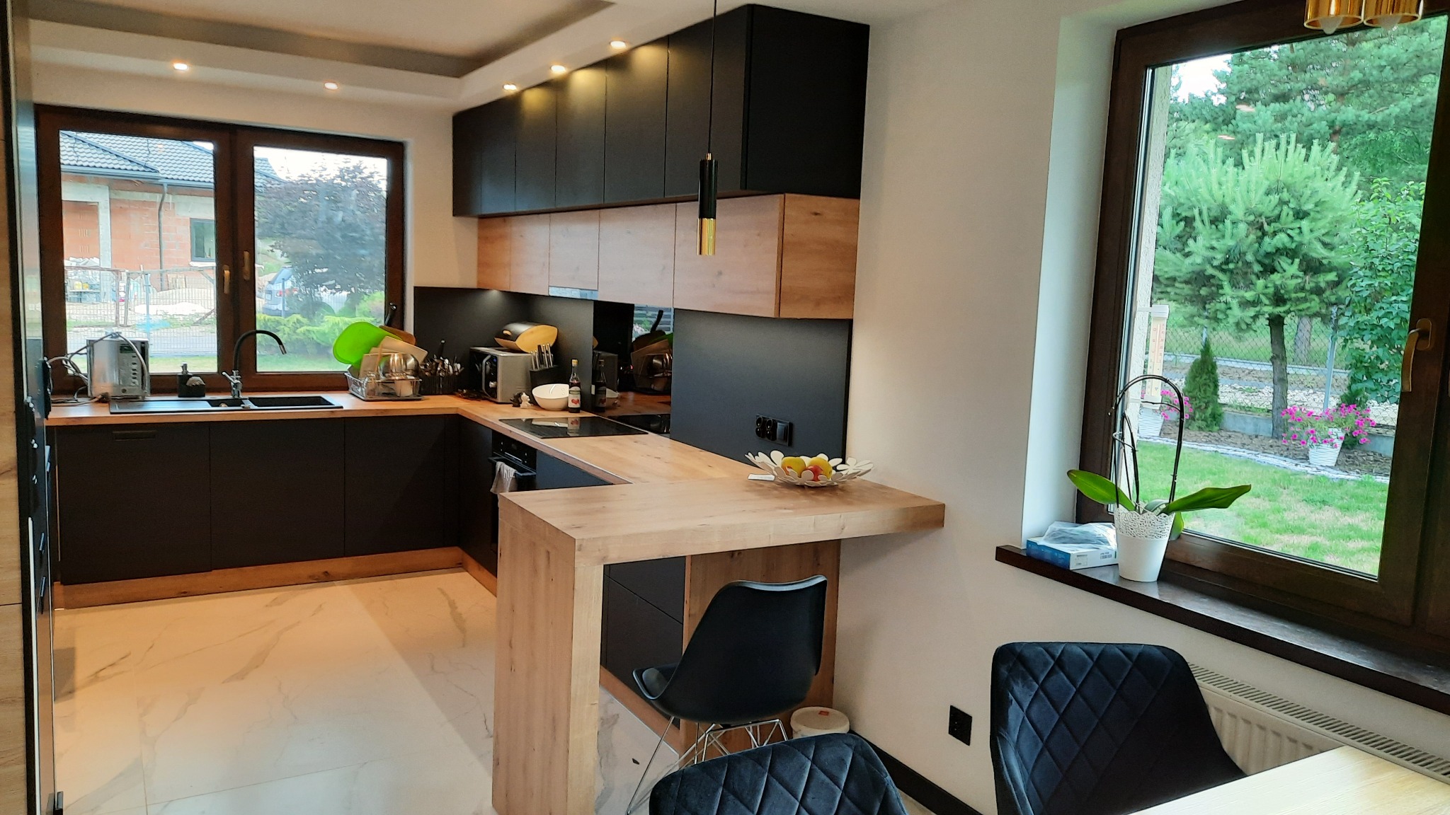 Nowoczesna kuchnia z czarnymi szafkami i drewnianym blatem, widok na ogród przez okno, marmurowe płytki podłogowe, czarne krzesła barowe.