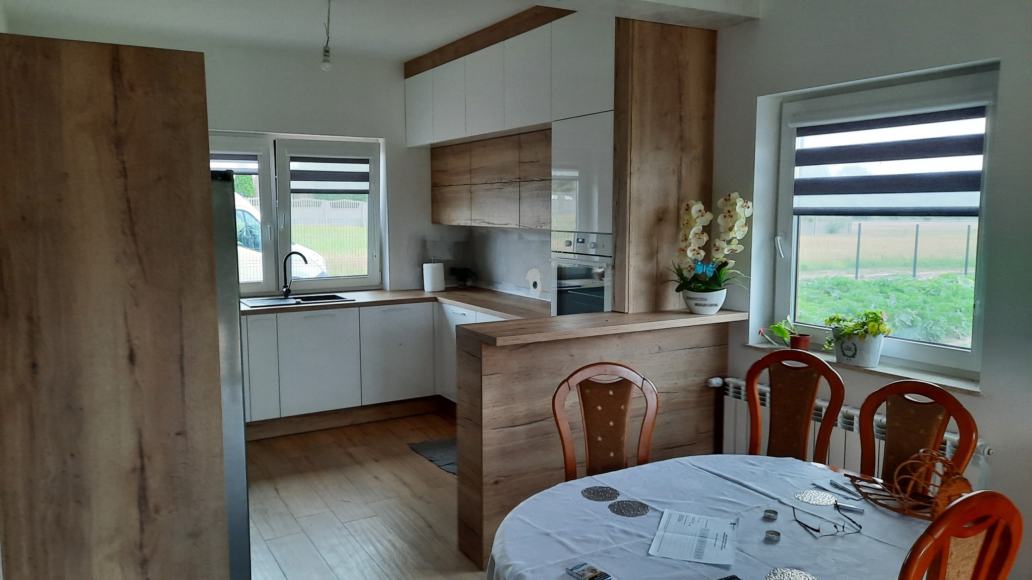 Jasna kuchnia z drewnianymi akcentami, białe szafki górne i dolne, drewniany blat, stół z krzesłami w pierwszym planie, widok na ogród przez okno.