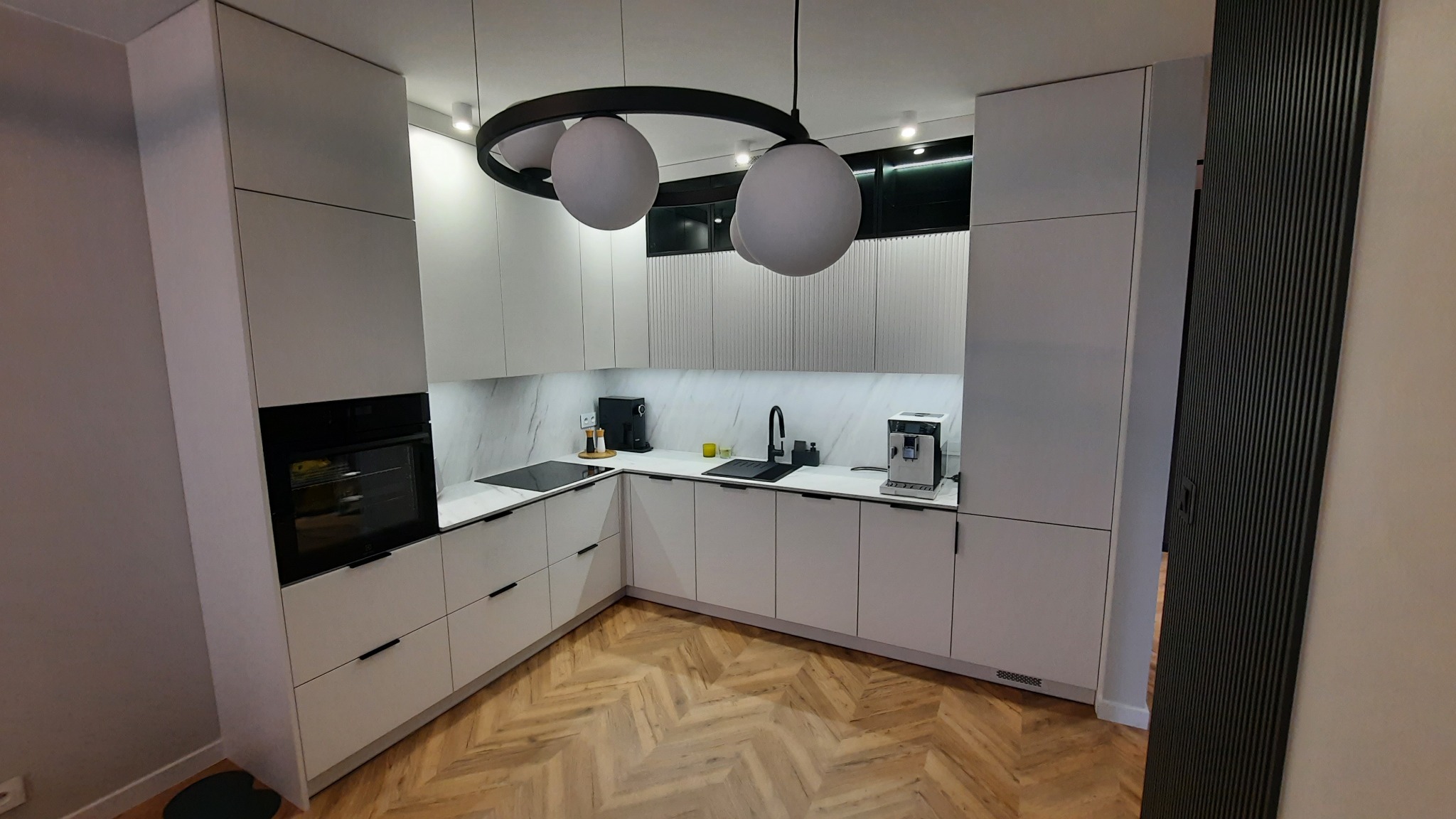 Nowoczesna kuchnia z białymi frontami, czarnymi akcentami i parkietem w jodełkę, oświetlona lampą z trzema kulami.