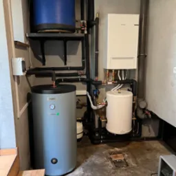 Zestaw urządzeń grzewczych w kotłowni: szary zbiornik Vasti z manometrem, biały zbiornik Galmet, biały piec Samsung, rury w izolacji, oraz fragment odkrytej podłogi z deską.