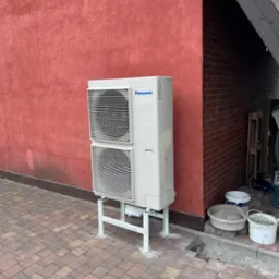 Zewnętrzna jednostka pompy ciepła Panasonic zamontowana na metalowym stelażu przy czerwonej ścianie budynku, widoczne również elementy kostki brukowej i wiaderka z zaprawą.