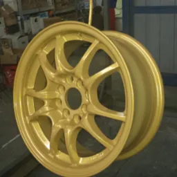 Dwa złote felgi aluminiowe po malowaniu proszkowym, wiszące na wieszaku w warsztacie.