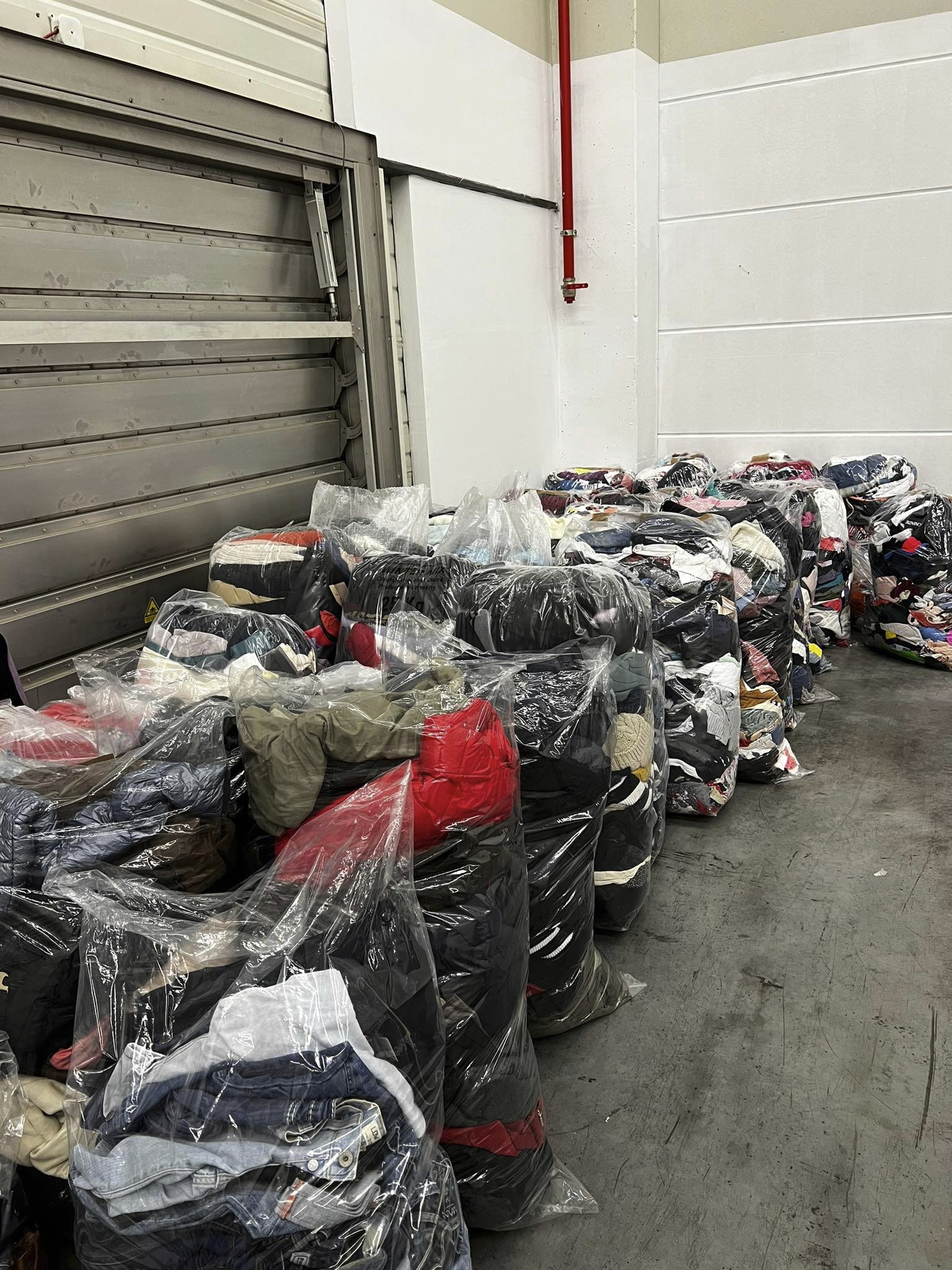 Magazyn z workami przepełnionymi odzieżą, zapakowanymi w przezroczyste folie, widoczne zróżnicowane ubrania wewnątrz, szara podłoga i ściany magazynowe w tle.