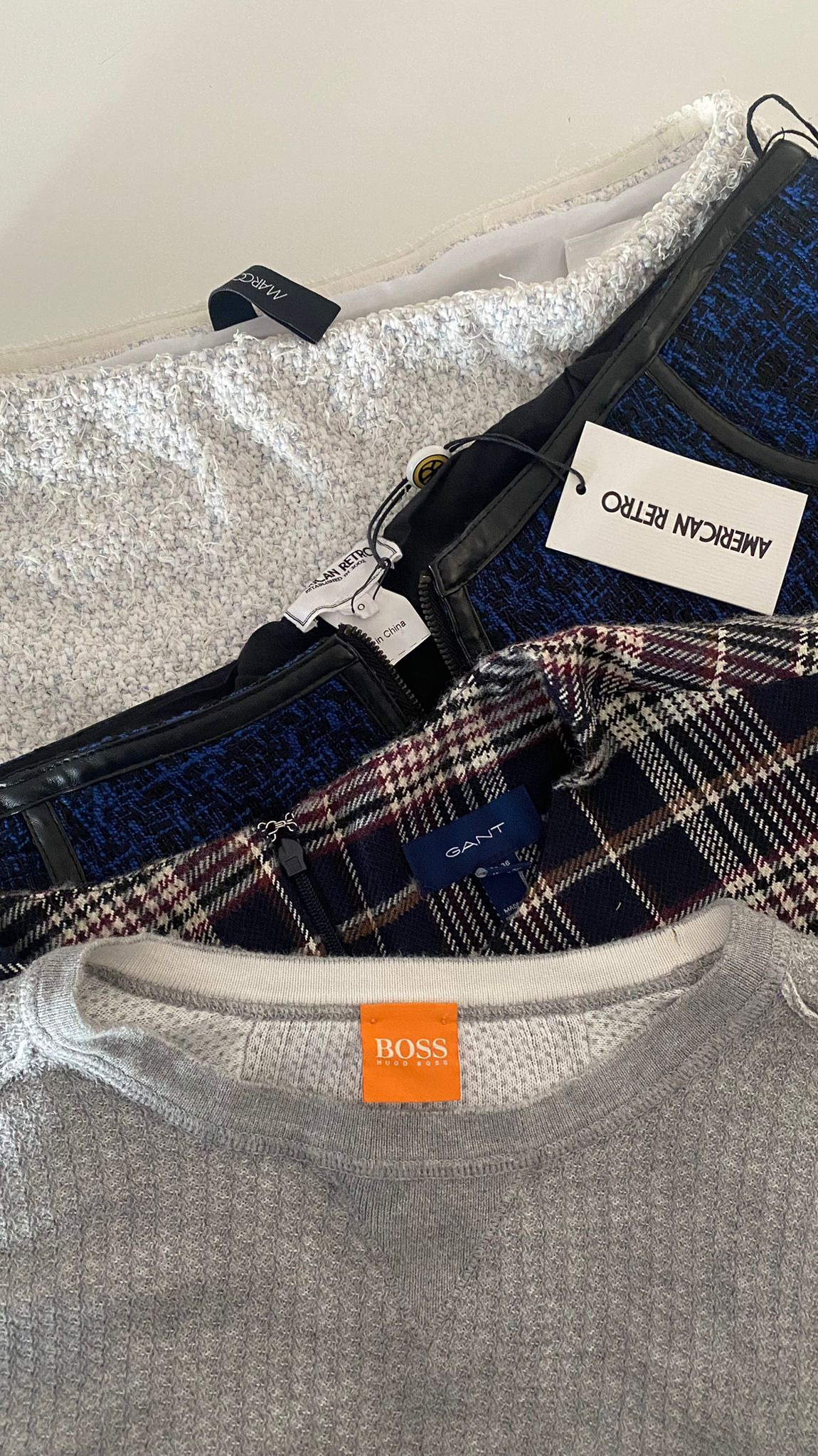 Stos ubrań różnych marek: biała spódnica z cekinami, niebieska spódnica w kratę z czarną skórą, spódnica w kratę Gant i szara bluza Hugo Boss, prezentowane we Wrocławiu.