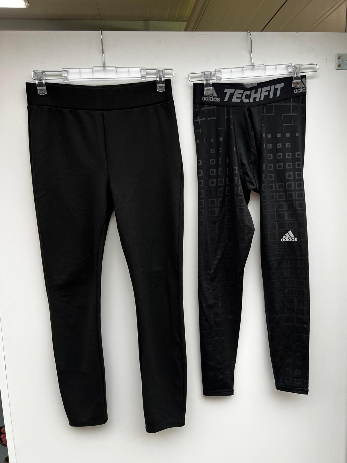 Dwie pary czarnych legginsów sportowych wiszące na wieszakach: gładkie legginsy po lewej i legginsy Adidas Techfit z geometrycznym wzorem po prawej.