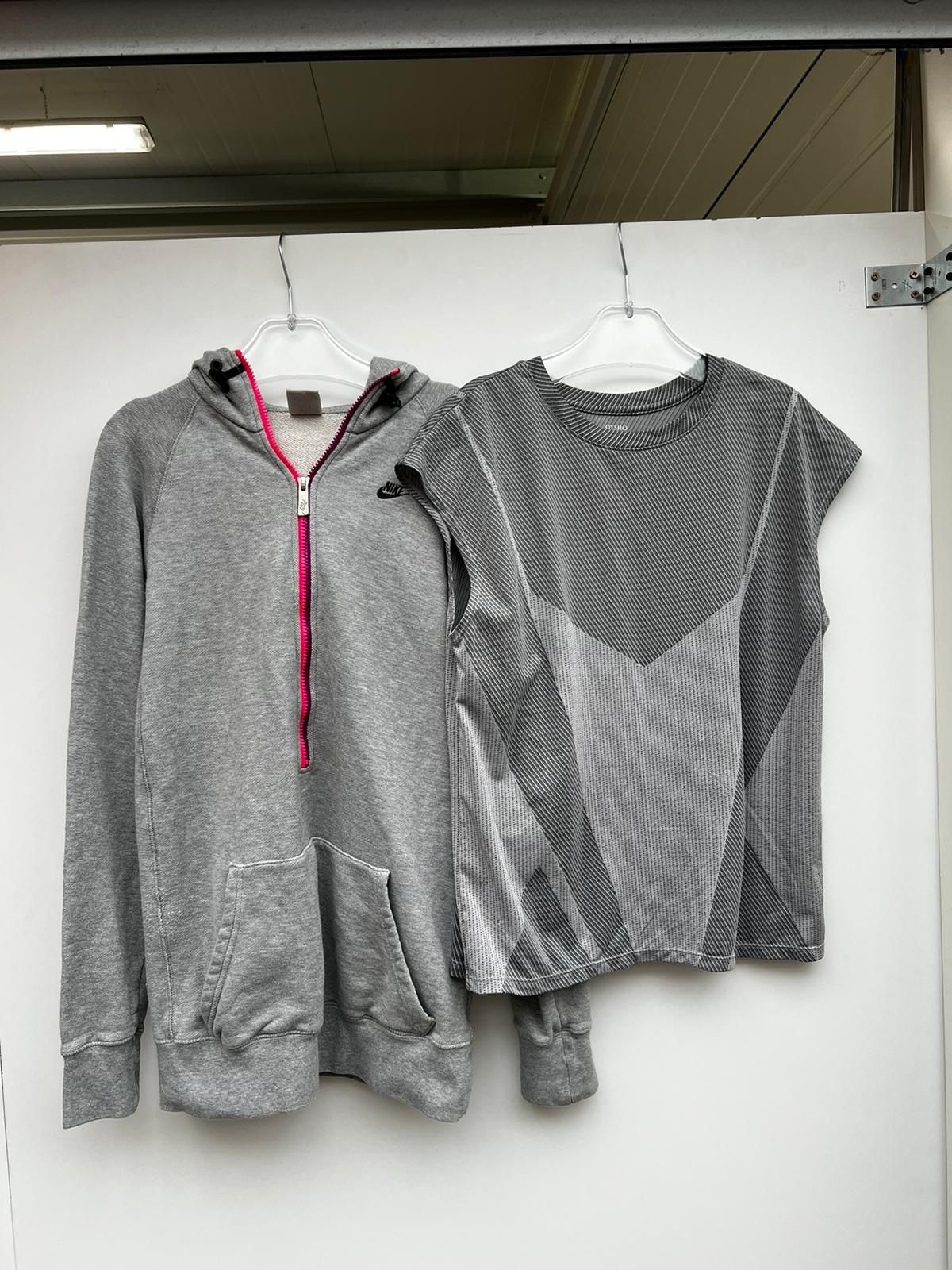 Dwie szare sportowe ubrania wiszące na wieszakach: bluza z kapturem z różowym zamkiem i logo Nike oraz bezrękawnik z geometrycznym wzorem.