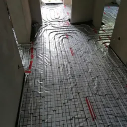 Białe rury ogrzewania podłogowego ułożone na czarnej siatce z czerwonymi elementami mocującymi, widoczne w surowym wnętrzu budynku w trakcie instalacji.