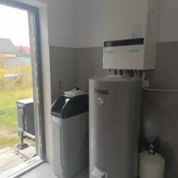 Nowoczesna kotłownia: biały piec gazowy Panasonic, szary zasobnik Ferroli i stacja uzdatniania wody w jasnym pomieszczeniu z szarymi płytkami i oknem.