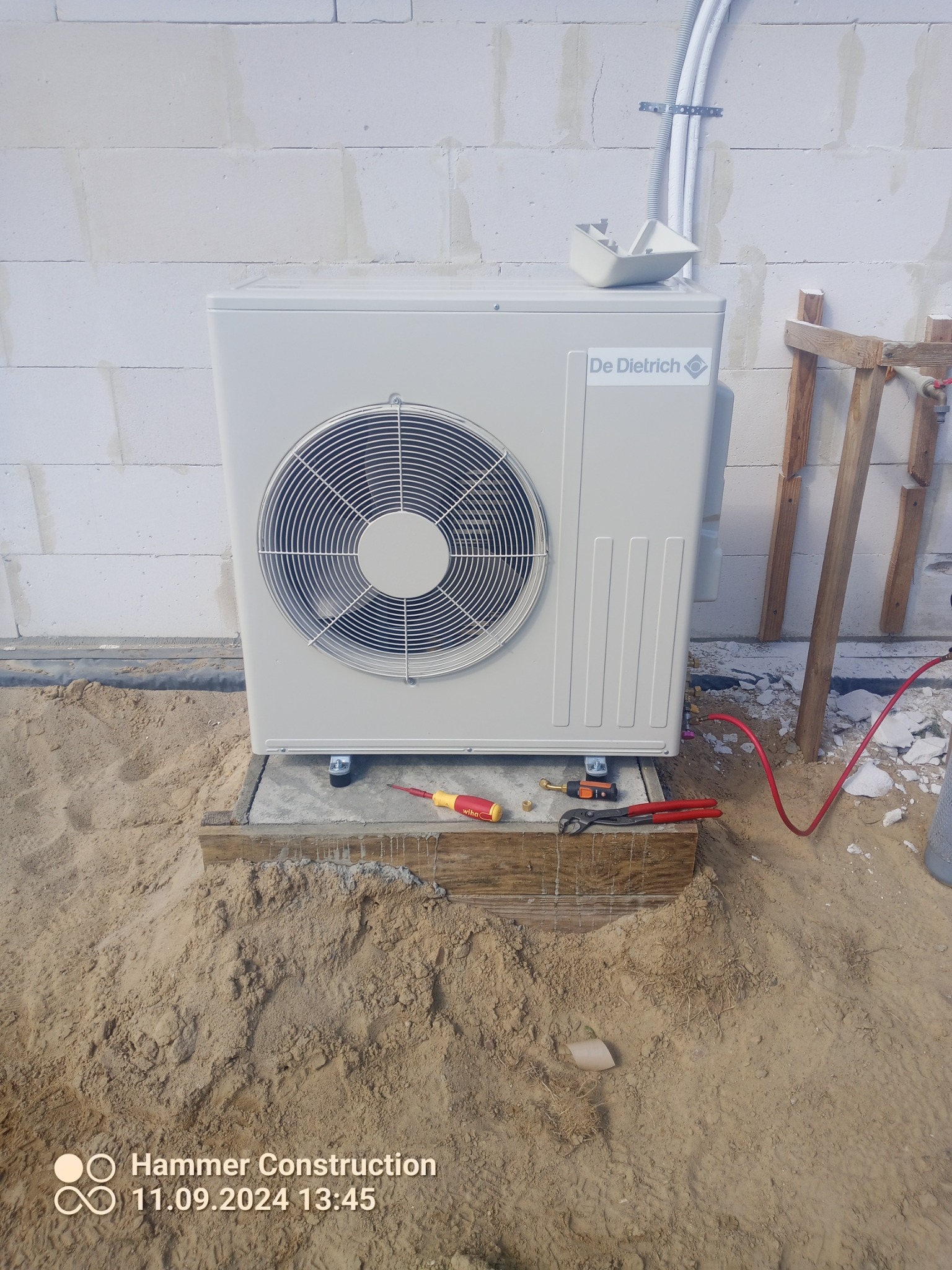 Zewnętrzna jednostka pompy ciepła De Dietrich na betonowej podstawie, z widocznymi narzędziami (wkrętak, kombinerki) i fragmentem instalacji, na tle ściany z bloczków betonowych w trakcie montażu.