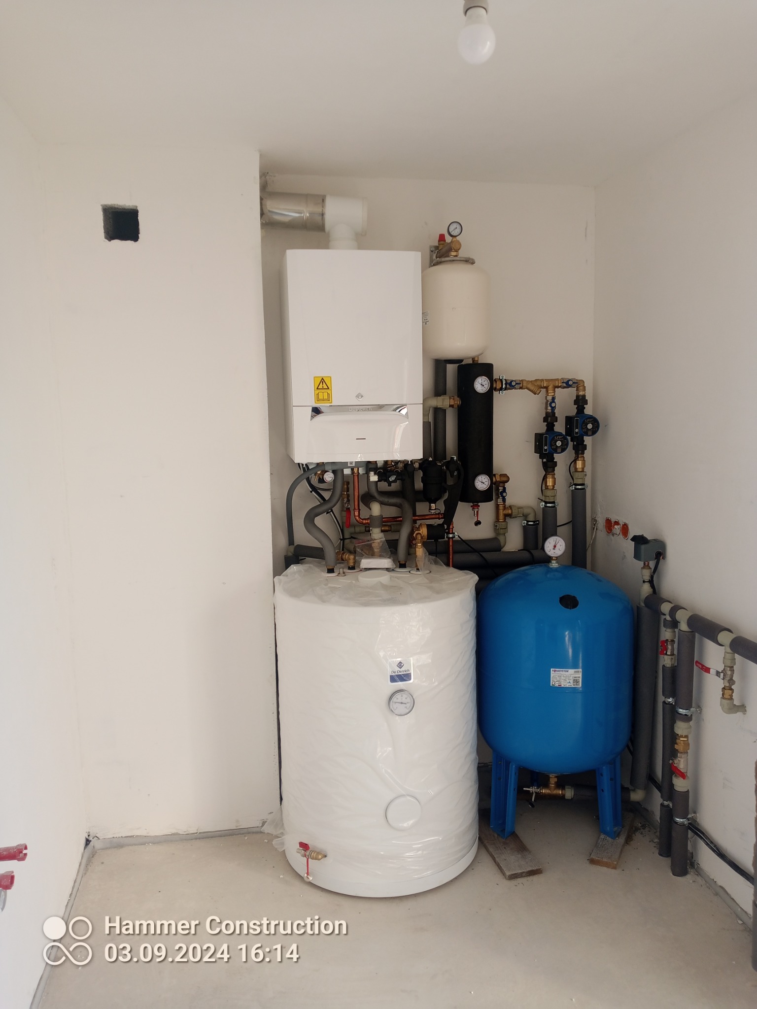 Nowa instalacja grzewcza w kotłowni: piec gazowy De Dietrich, biały zasobnik ciepłej wody owinięty folią ochronną, niebieskie naczynie przeponowe Aquasystem na drewnianych podkładkach, rury...