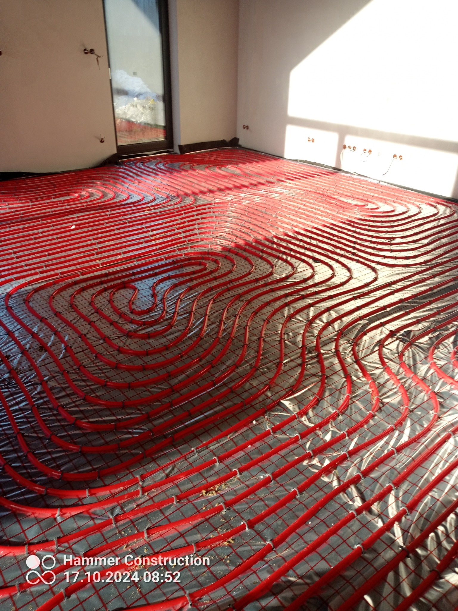 Czerwone rury ogrzewania podłogowego ułożone spiralnie na srebrnej folii izolacyjnej w jasnym pomieszczeniu z oknem, widoczne uchwyty mocujące rury do podłoża.