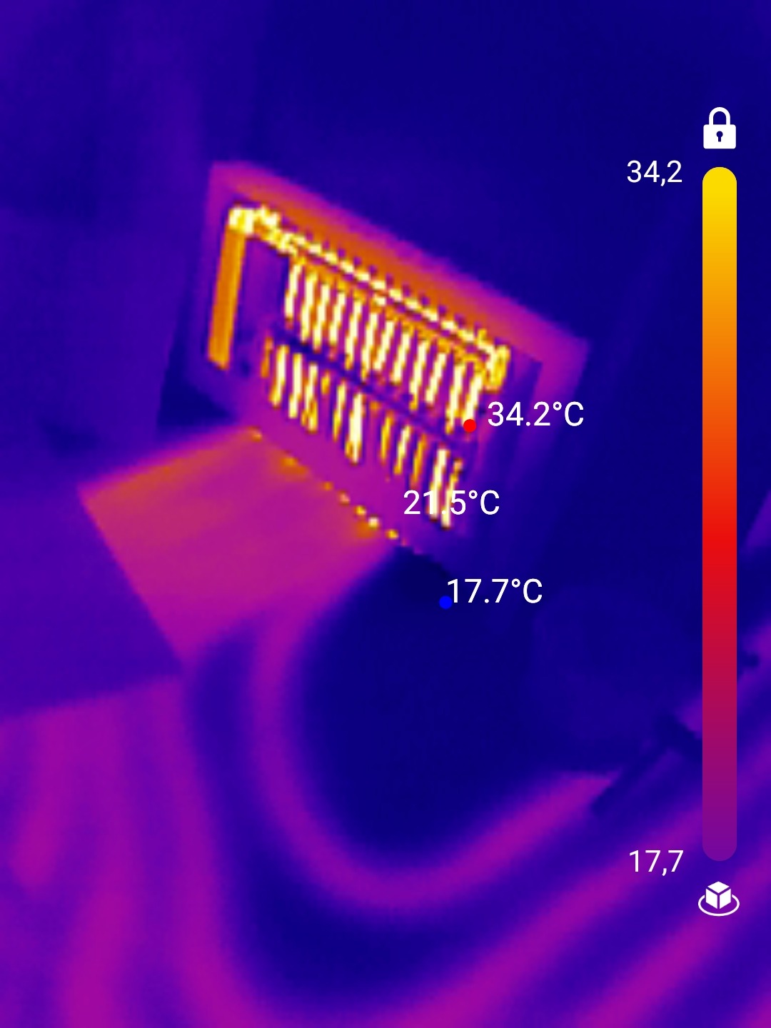 Termowizyjne zdjęcie grzejnika, ukazujące rozkład temperatur na żeberkach, z zaznaczonymi punktami pomiarowymi 34.2°C, 21.5°C i 17.7°C, na tle podłogi z widocznym wzorem.