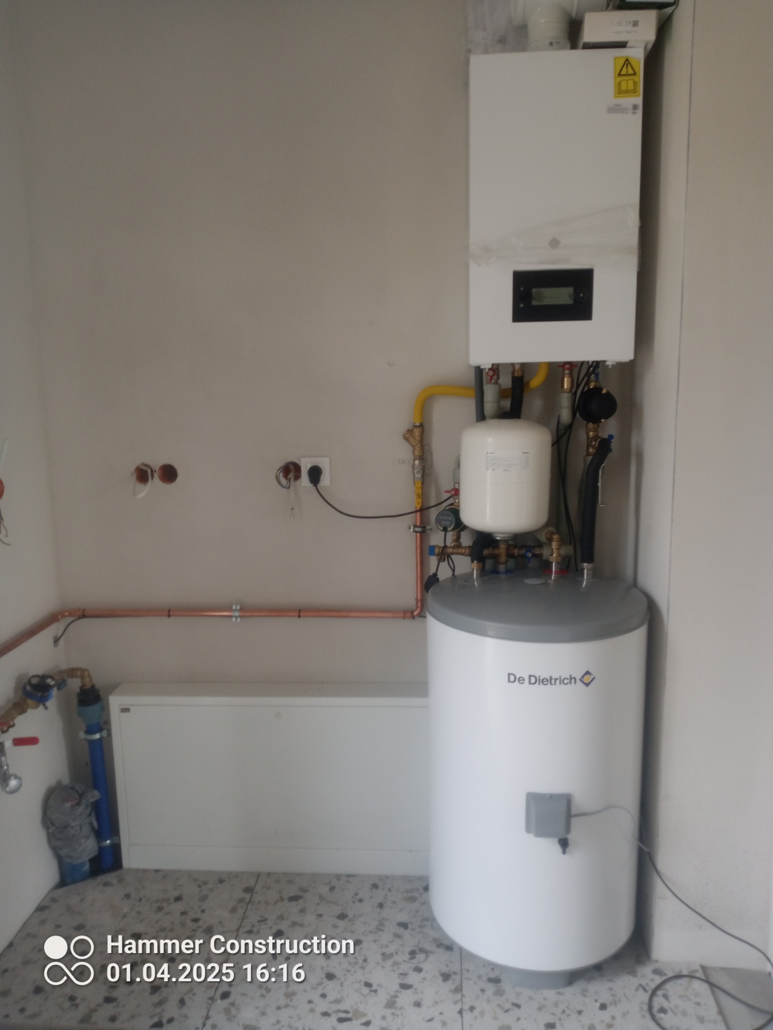 Biały piec gazowy De Dietrich zamontowany na ścianie w pomieszczeniu z widocznymi rurami miedzianymi i instalacją elektryczną.