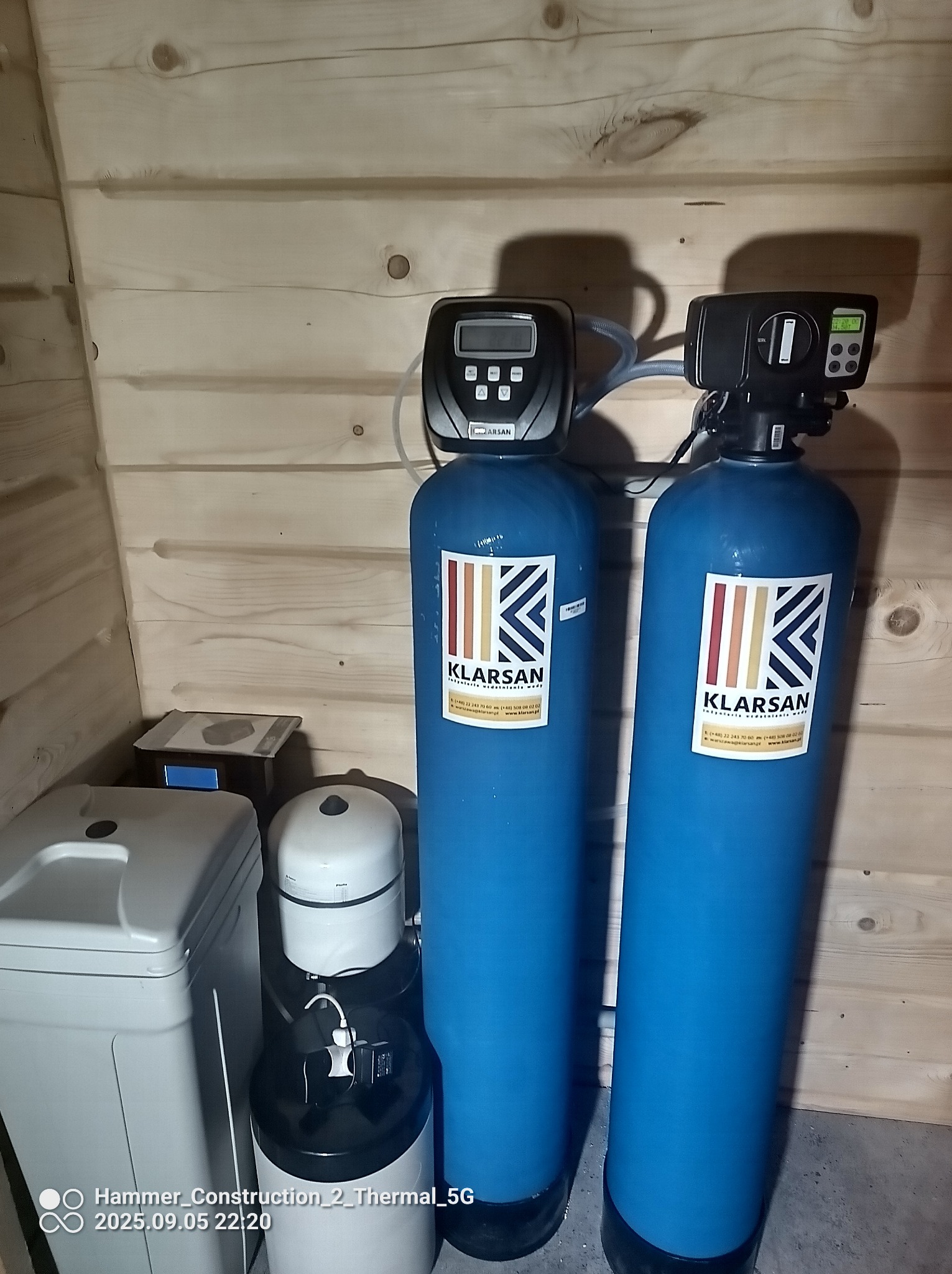 Dwa niebieskie filtry do wody marki Klarsan w pomieszczeniu wyłożonym jasnym drewnem. Widoczny zbiornik na sól i inne elementy instalacji.