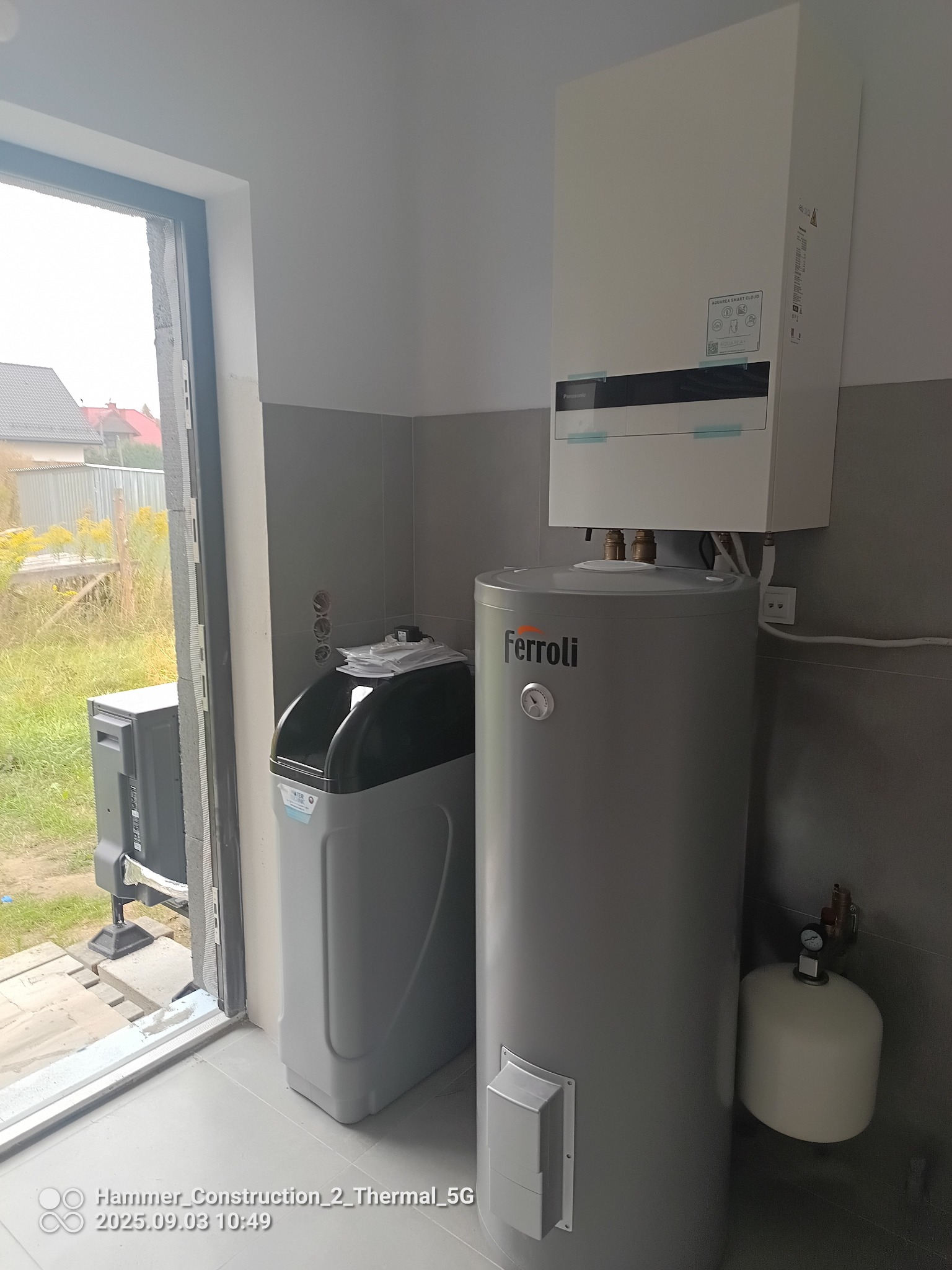 Nowoczesna kotłownia: biały piec gazowy Panasonic, szary zasobnik Ferroli i stacja uzdatniania wody w jasnym pomieszczeniu z szarymi płytkami i oknem.