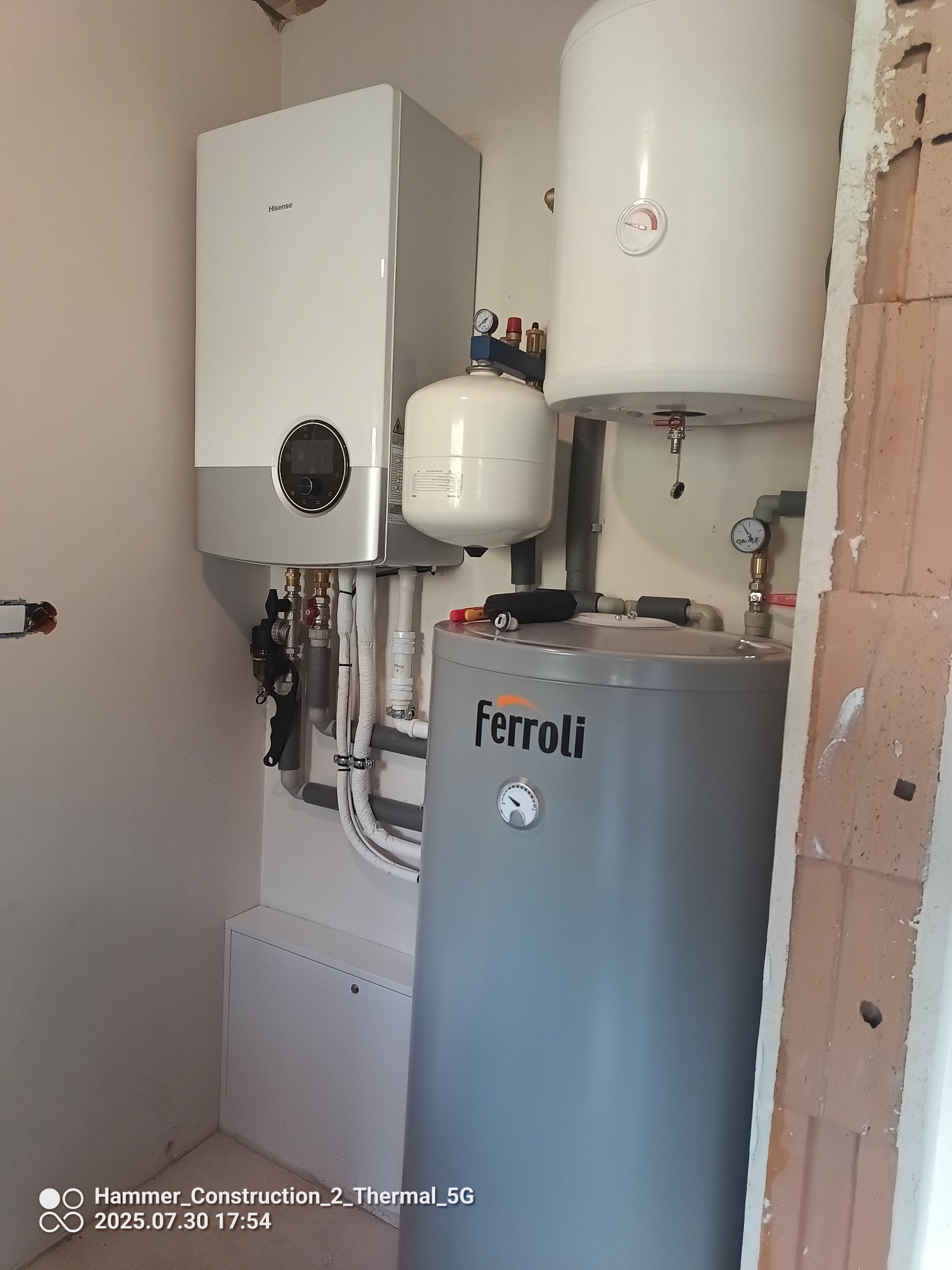 Nowoczesny piec gazowy Hisense i zasobnik Ferroli, instalacja grzewcza z rurami i zaworami, widok z bliska na kotłownię w trakcie prac wykończeniowych.