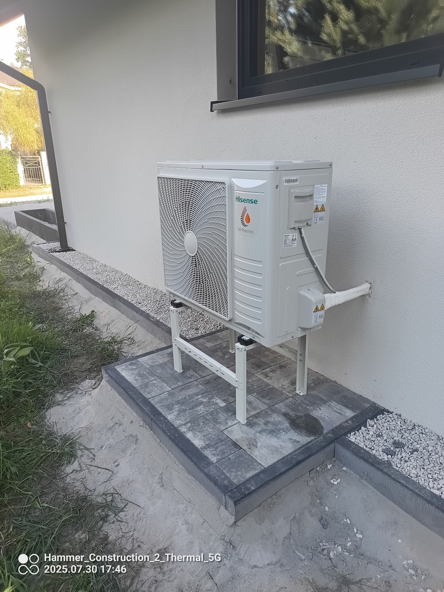 Biała pompa ciepła Hisense Hi-Therma, zamontowana na zewnątrz budynku na metalowym stelażu, z widocznymi przewodami w izolacji wchodzącymi w ścianę.