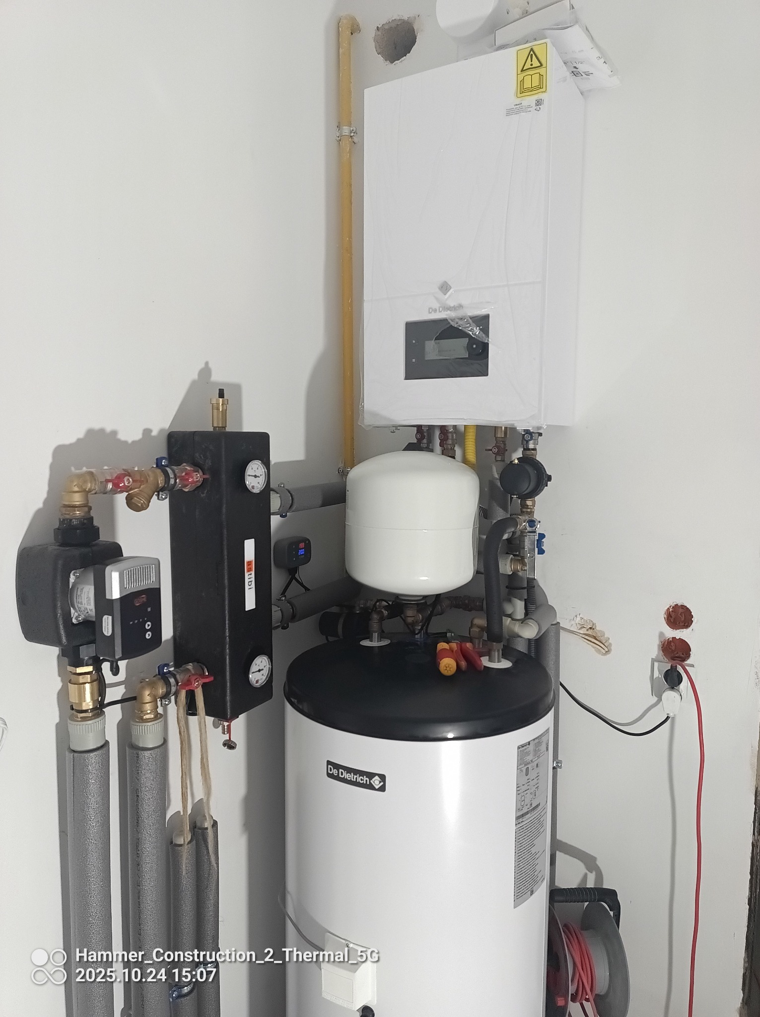 Biały piec gazowy De Dietrich z podłączonymi rurami i osprzętem, zamontowany na ścianie w pomieszczeniu. Widoczne elementy instalacji i podłączenia elektryczne.