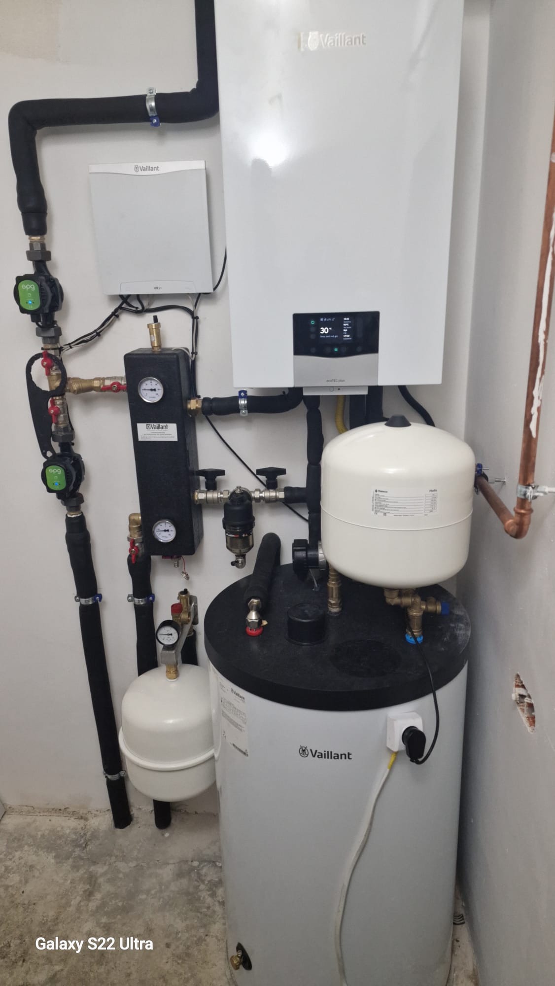 Biały piec gazowy Vaillant z czarnymi rurami i manometrami na ścianie w kotłowni. Widoczny zbiornik wyrównawczy i elementy instalacji.