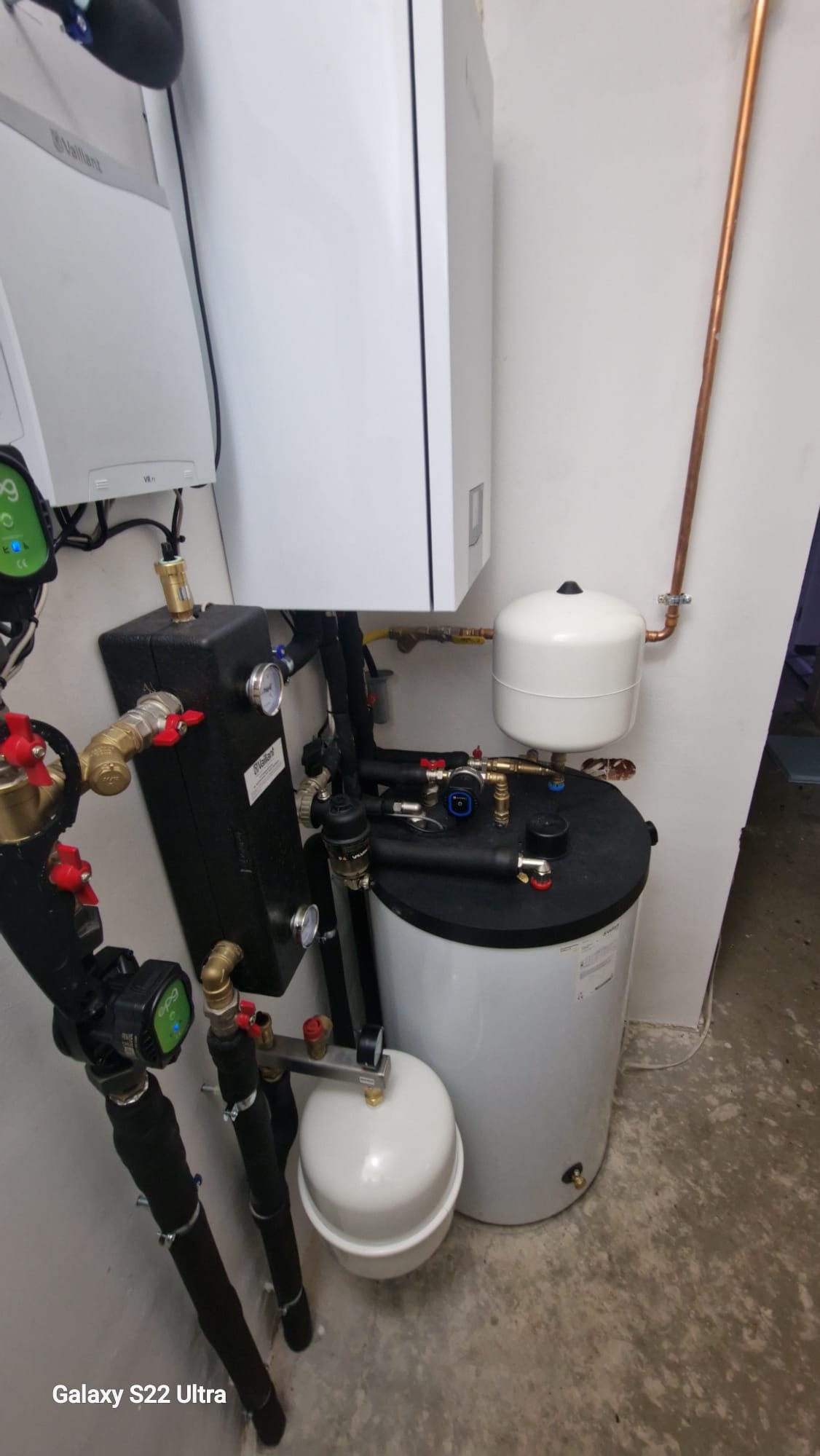 Złożona instalacja grzewcza z piecem Vaillant, zbiornikiem buforowym i pompami obiegowymi, widoczna w kotłowni. Rury miedziane i czarne, manometry, zawory.