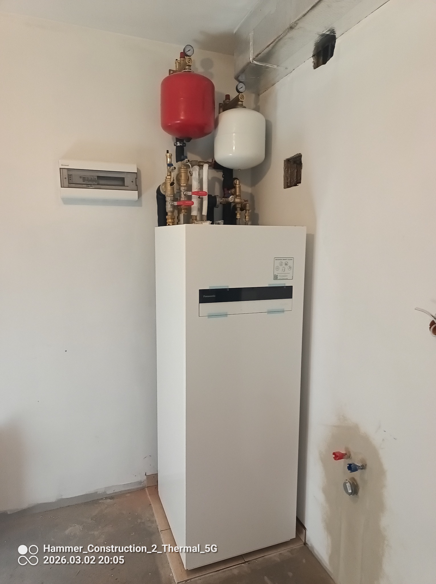 Biały piec gazowy Panasonic z czerwonym i białym zbiornikiem wyrównawczym, widoczne rury i zawory. Ściany w trakcie wykończenia, widoczne ślady po instalacji.