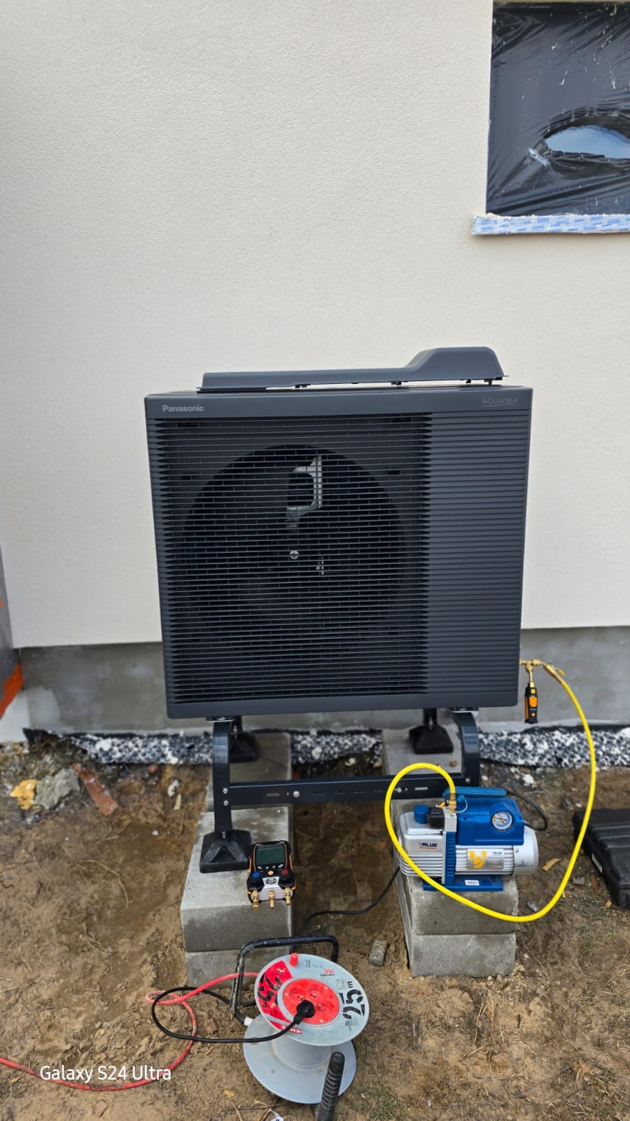 Czarna pompa ciepła Panasonic Aquarea zamontowana na betonowych blokach, podłączona do urządzeń pomiarowych i pompy próżniowej. Okno zabezpieczone folią w tle.