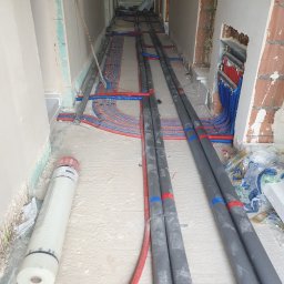 USŁUGI HYDRAULICZNE I BUDOWLANE AGNIESZKA ŚLIWIŃSKA-ZAWIŚLAK - Korytarz w trakcie instalacji ogrzewania podłogowego i rur wodnych, widoczne rury grzewcze w izolacji, rozdzielacz na ścianie i rolka siatki do wylewki.