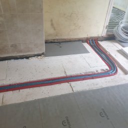USŁUGI HYDRAULICZNE I BUDOWLANE AGNIESZKA ŚLIWIŃSKA-ZAWIŚLAK - Instalacja ogrzewania podłogowego: rury grzewcze w czerwonej otulinie ułożone na styropianie z nadrukiem PURMO, widoczne fragmenty podłoża i ściany w trakcie prac wykończeniowych.