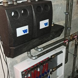 USŁUGI HYDRAULICZNE I BUDOWLANE AGNIESZKA ŚLIWIŃSKA-ZAWIŚLAK - Rozdzielacz ogrzewania podłogowego z rurami w kolorze niebieskim i czerwonym, zamontowany na ścianie z instalacją hydrauliczną i zbiornikiem buforowym z logo producenta.