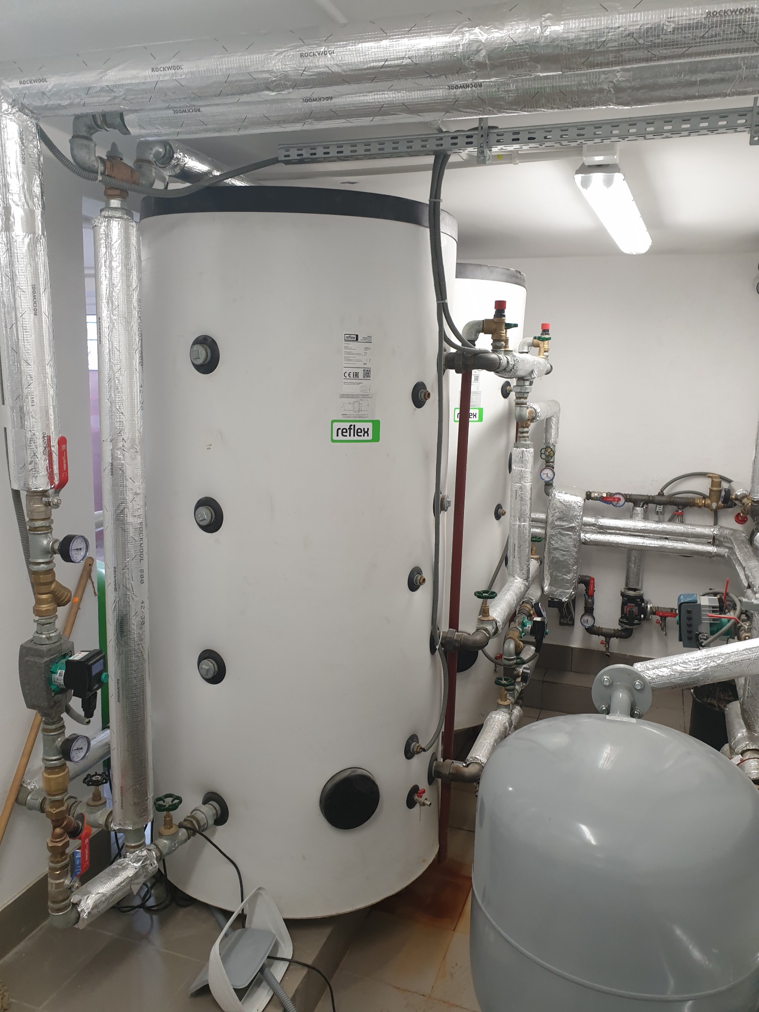 Dwa białe, cylindryczne zbiorniki z licznymi przyłączami rur i zaworów, zaizolowane rury Rockwool na ścianie i suficie, szara butla ciśnieniowa i biały skaner na podłodze.