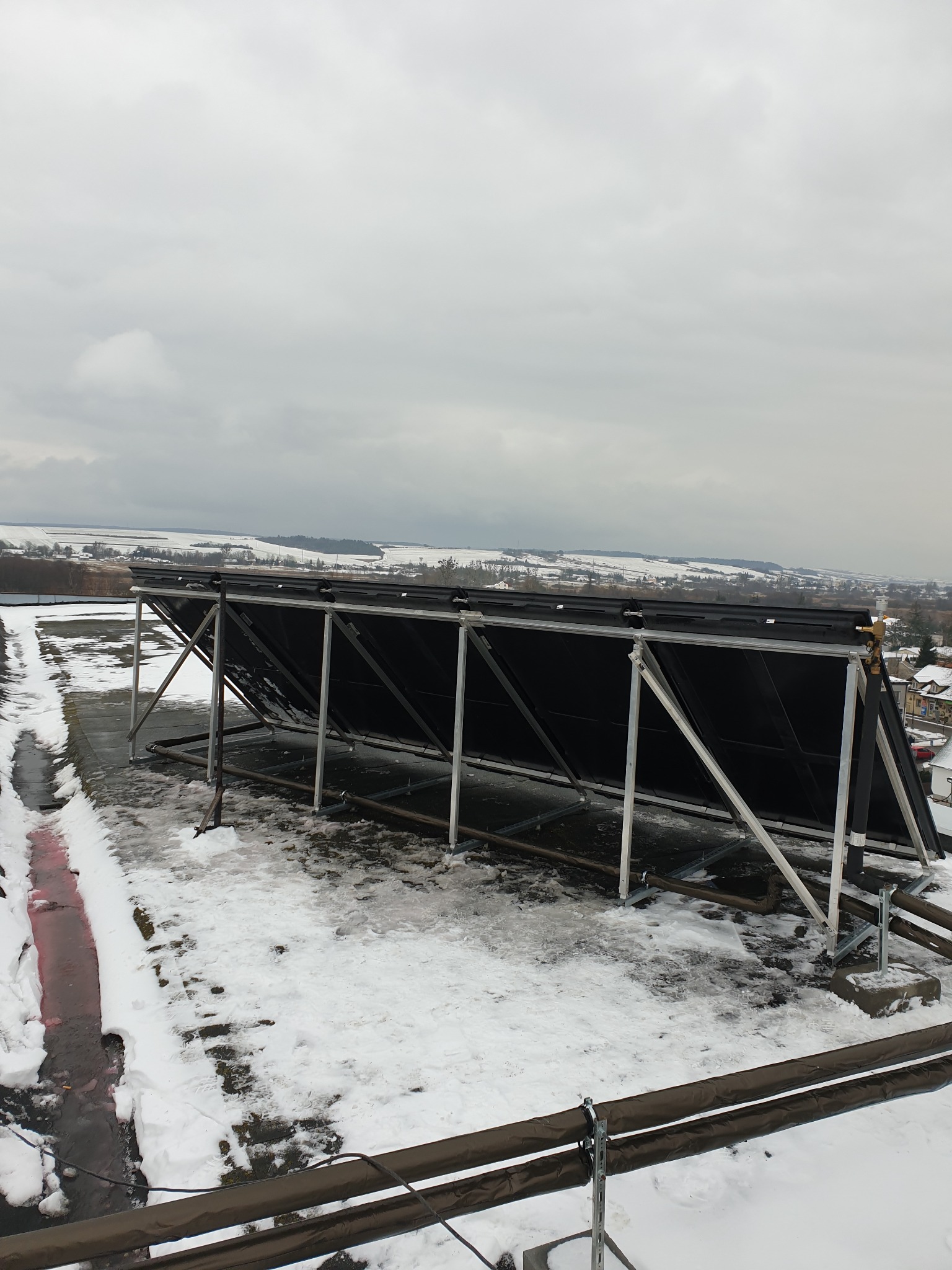 Czarne panele kolektorów słonecznych na metalowej konstrukcji, zamontowane na płaskim dachu pokrytym śniegiem, z widokiem na zimowy krajobraz w tle.