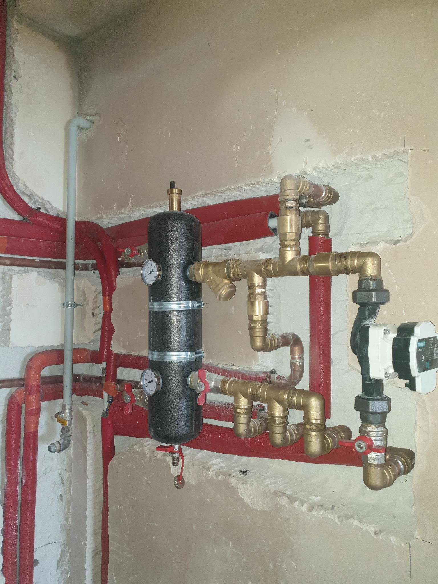 Złożona instalacja grzewcza z czerwonymi rurami, czarnym zbiornikiem buforowym z manometrami i mosiężnymi złączkami, widoczna pompa obiegowa, instalacja w surowym stanie, ściany z ubytkami tynku.