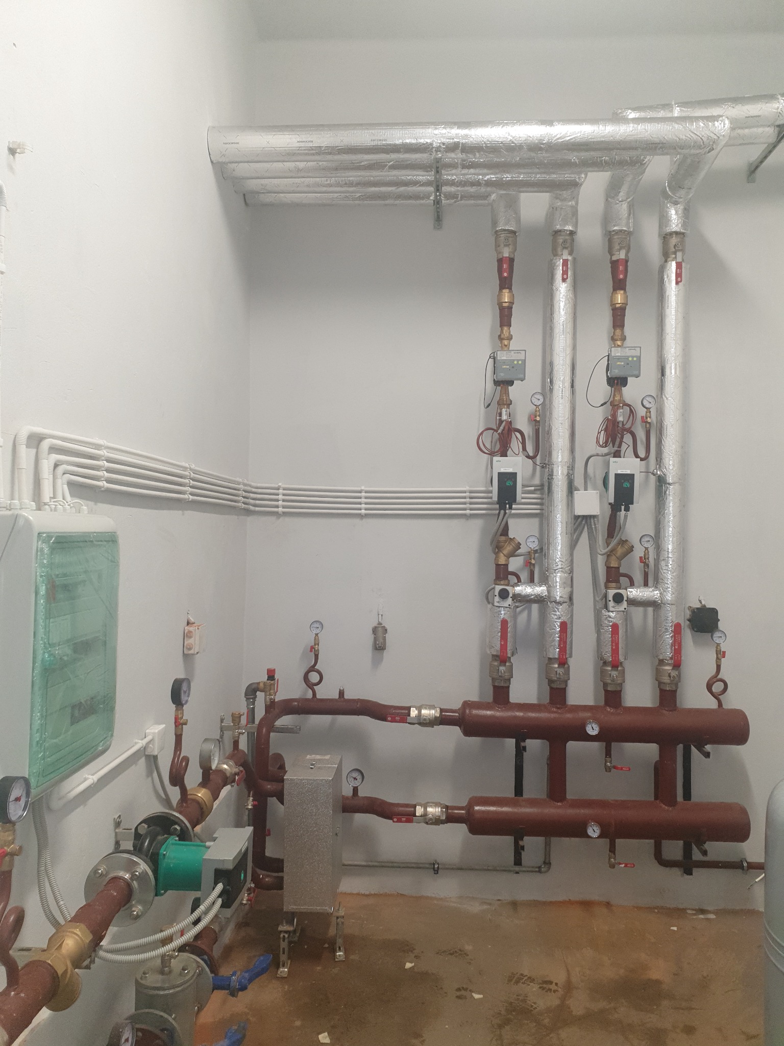 Instalacja z rur miedzianych i stalowych, zaworów, manometrów oraz izolowanych rur na ścianie w pomieszczeniu technicznym. Widoczny zielony obieg pompy, szare przewody i skrzynka elektryczna.
