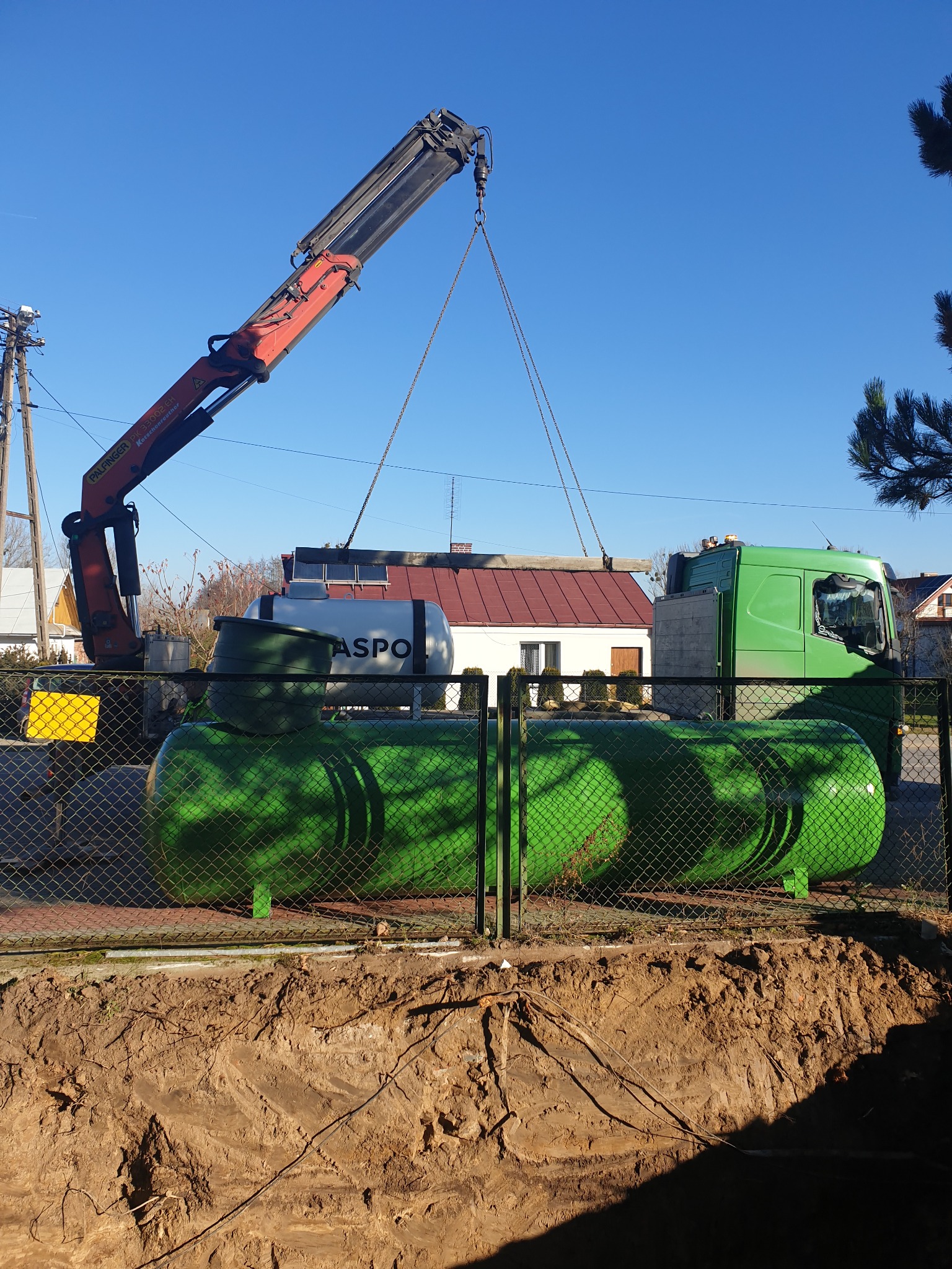Żuraw przenosi zielony zbiornik na gaz do wykopu, za ogrodzeniem widoczny biały zbiornik z napisem 'ASPO'.