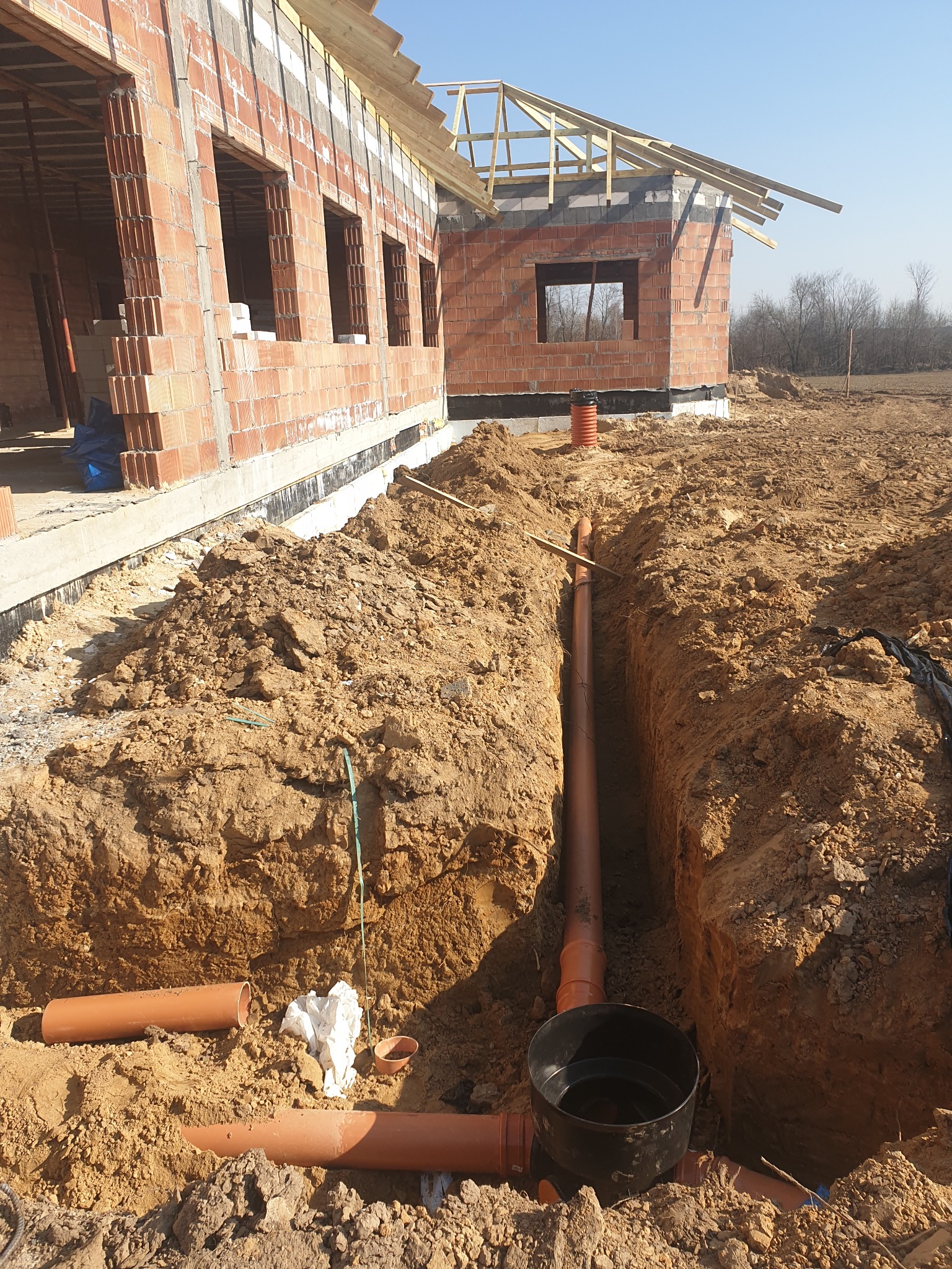 Wykop z ułożonymi rurami kanalizacyjnymi z tworzywa sztucznego, widok na budynek w stanie surowym otwartym z czerwonej cegły i drewnianą konstrukcją dachu, ziemisty teren budowy.
