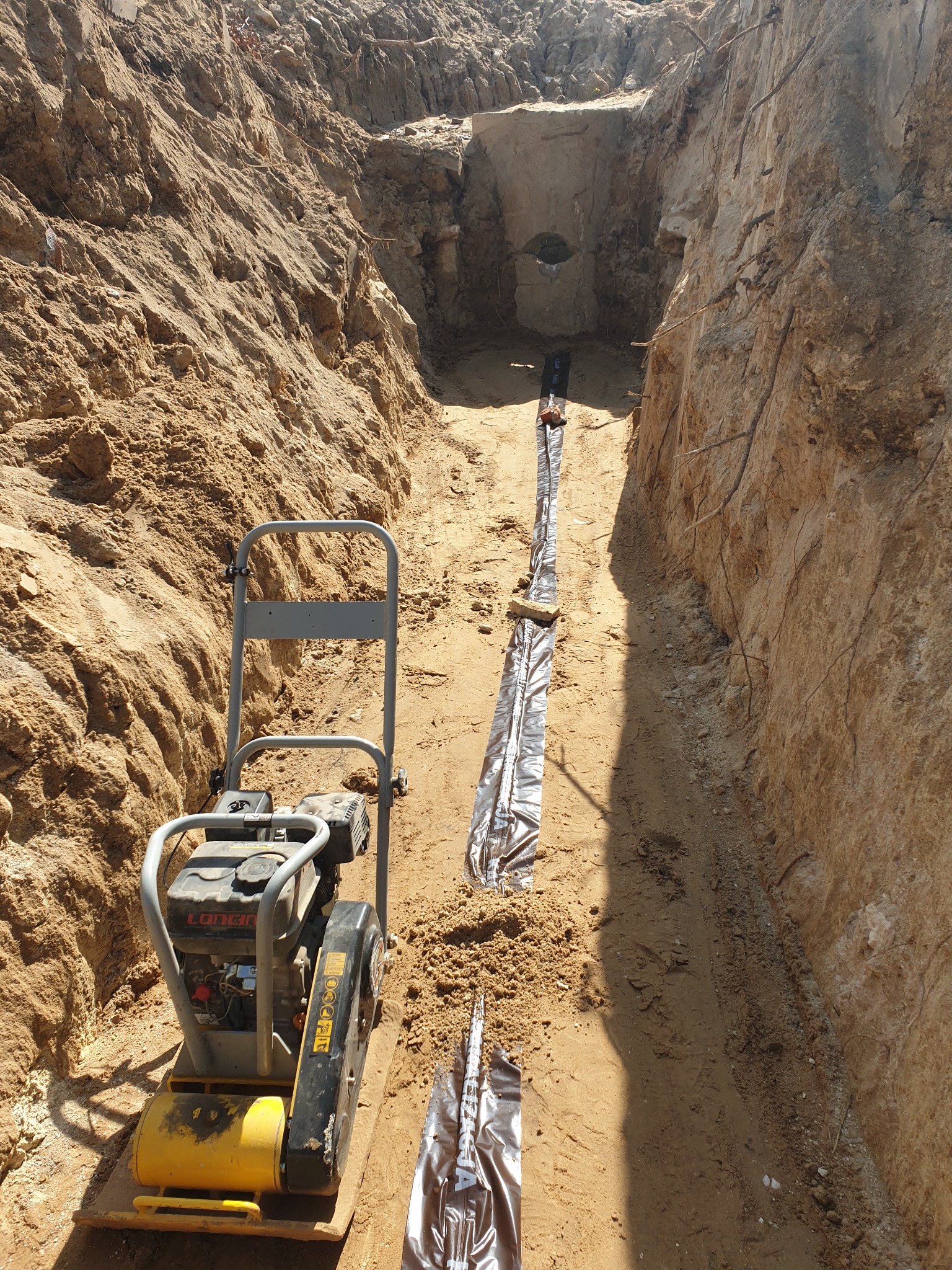 Głęboki wykop w ziemi z ułożoną rurą w folii oraz ubijarką wibracyjną na pierwszym planie, widoczne ślady prac ziemnych.