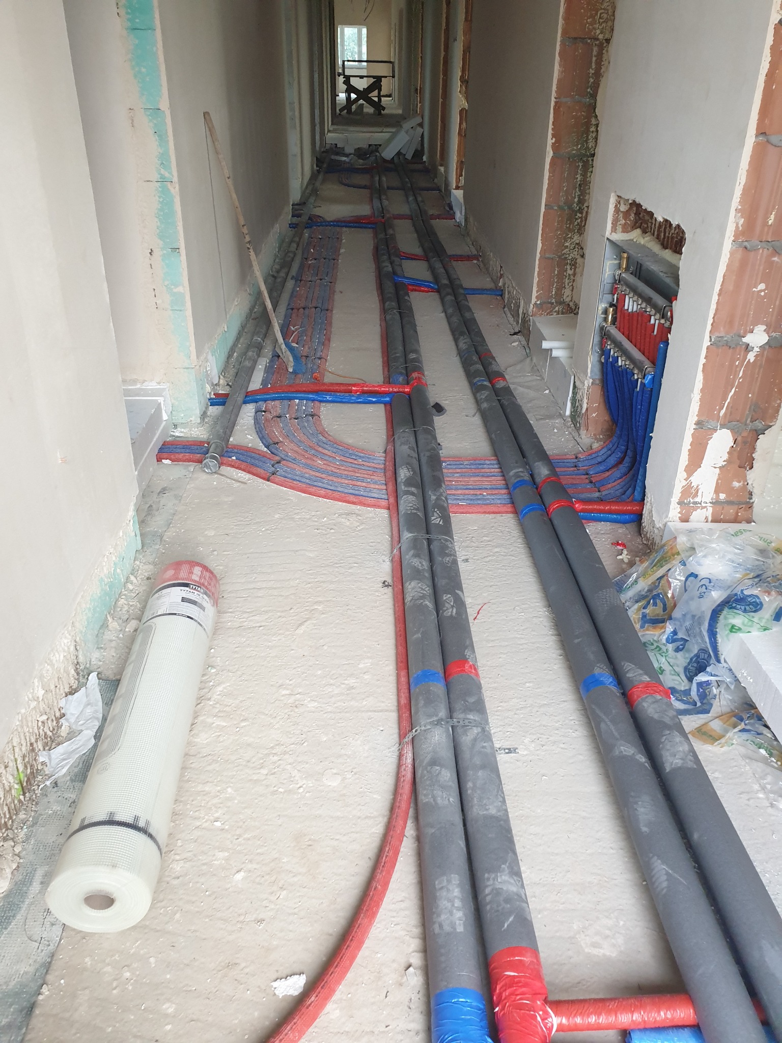 Korytarz w trakcie instalacji ogrzewania podłogowego i rur wodnych, widoczne rury grzewcze w izolacji, rozdzielacz na ścianie i rolka siatki do wylewki.