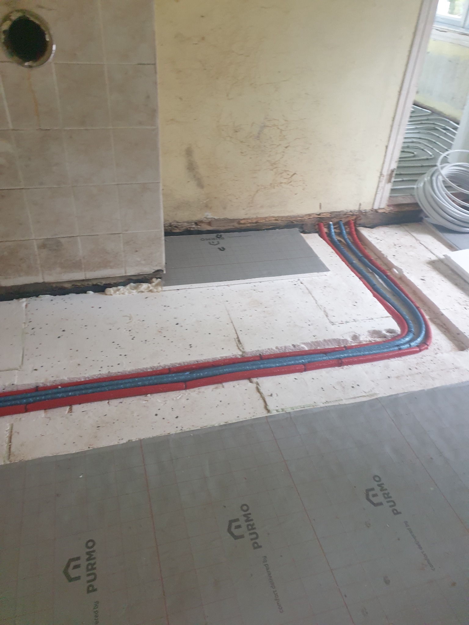 Instalacja ogrzewania podłogowego: rury grzewcze w czerwonej otulinie ułożone na styropianie z nadrukiem PURMO, widoczne fragmenty podłoża i ściany w trakcie prac wykończeniowych.