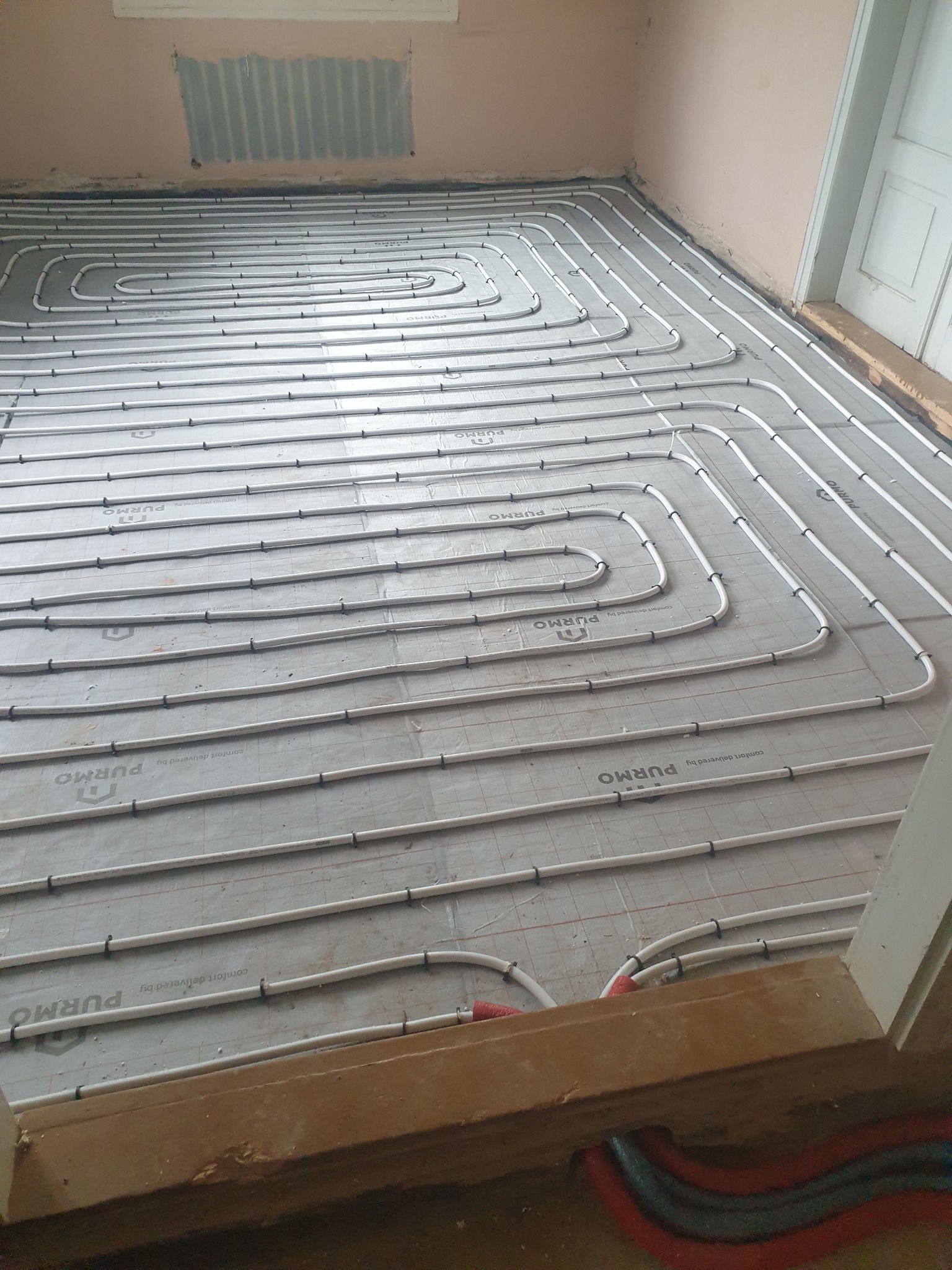Rozłożone białe rury na szarej macie z nadrukiem 'PURMO' tworzące system ogrzewania podłogowego w pomieszczeniu przed wylaniem posadzki.