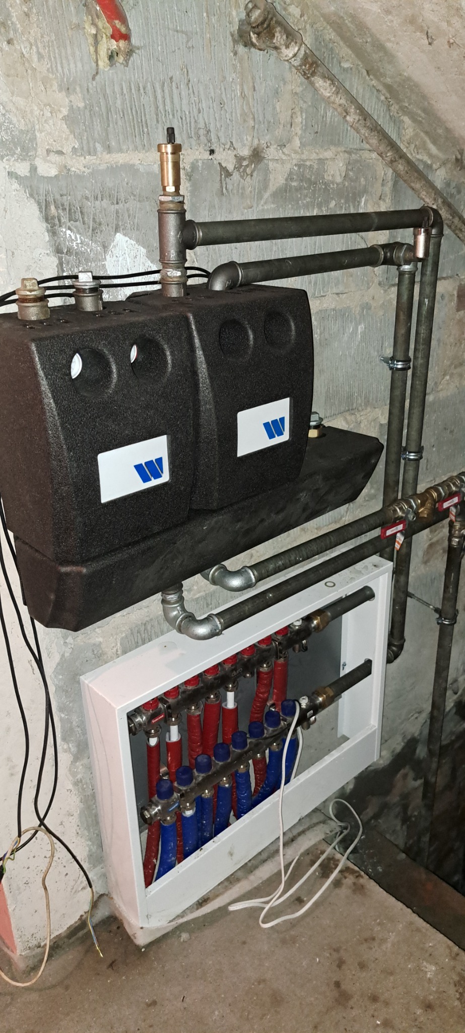 Rozdzielacz ogrzewania podłogowego z rurami w kolorze niebieskim i czerwonym, zamontowany na ścianie z instalacją hydrauliczną i zbiornikiem buforowym z logo producenta.