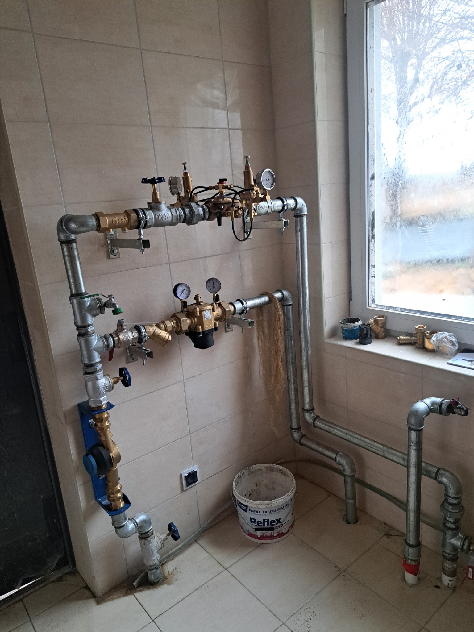 Kompleksowy system rur wodnych i gazowych z zaworami, manometrami i filtrami, zamontowany na kafelkowanej ścianie w pobliżu okna, widoczny również kubeł z farbą i różne złączki.