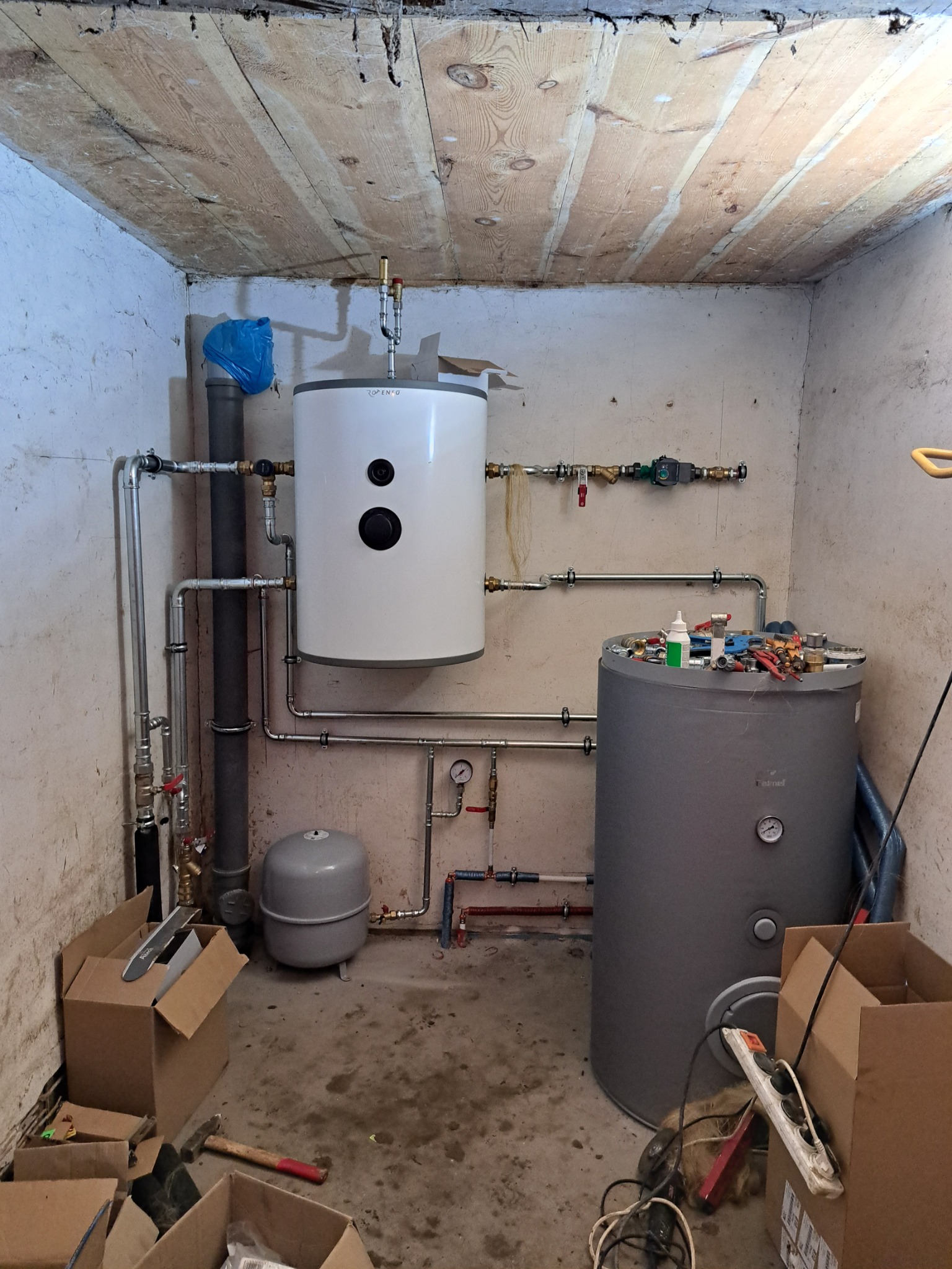 Dwa zbiorniki akumulacyjne w piwnicy, jeden biały, drugi szary, połączone rurami, widoczne narzędzia i kartony na podłodze, drewniany sufit.