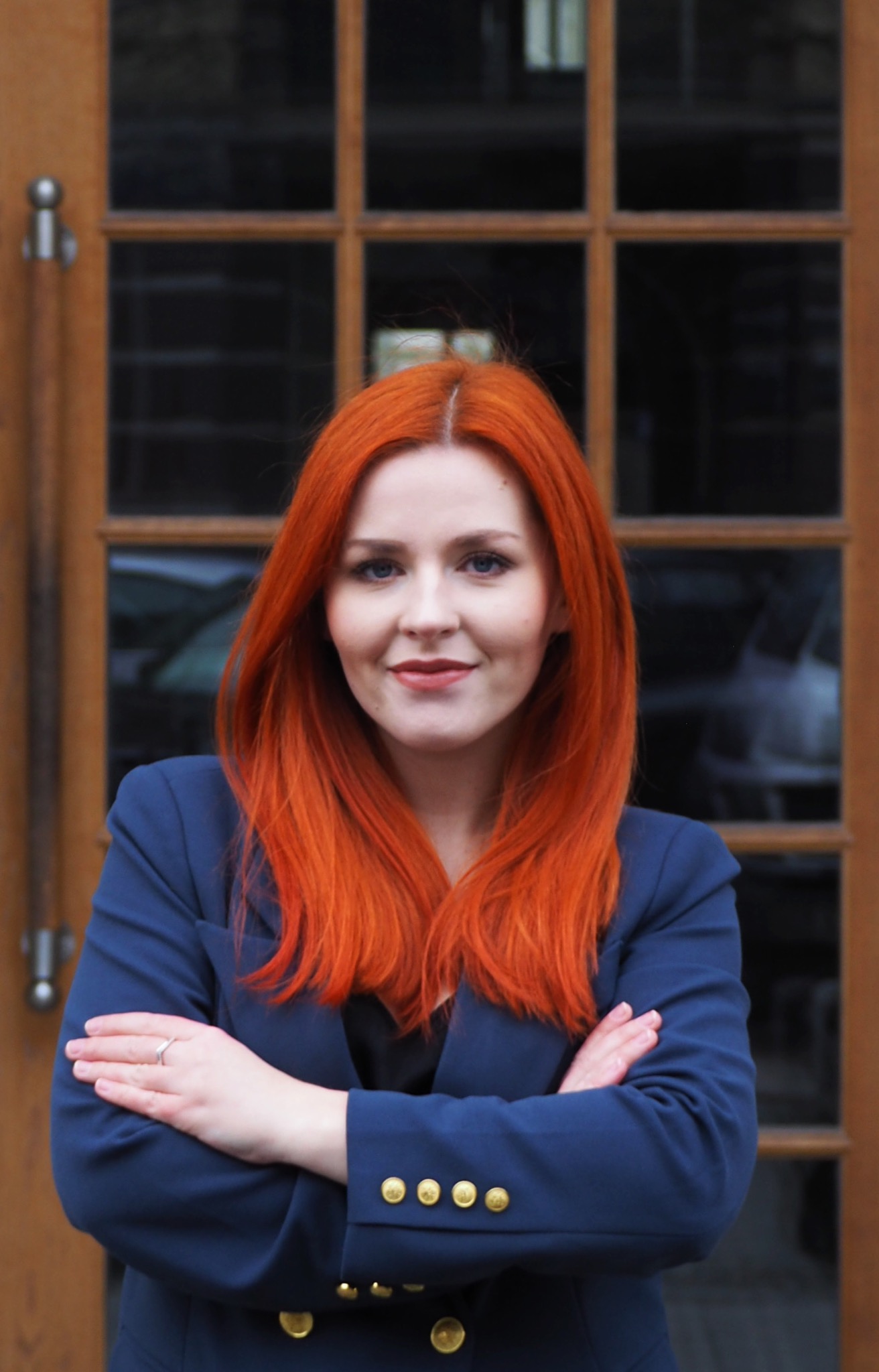 Portret kobiety o rudych włosach w granatowej marynarce z założonymi rękami na tle przeszklonych drzwi.