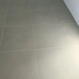 Szare, matowe płytki podłogowe o kwadratowym kształcie z jasnymi fugami, ułożone w regularną siatkę, przylegające do białych płytek ściennych w rogu pomieszczenia.